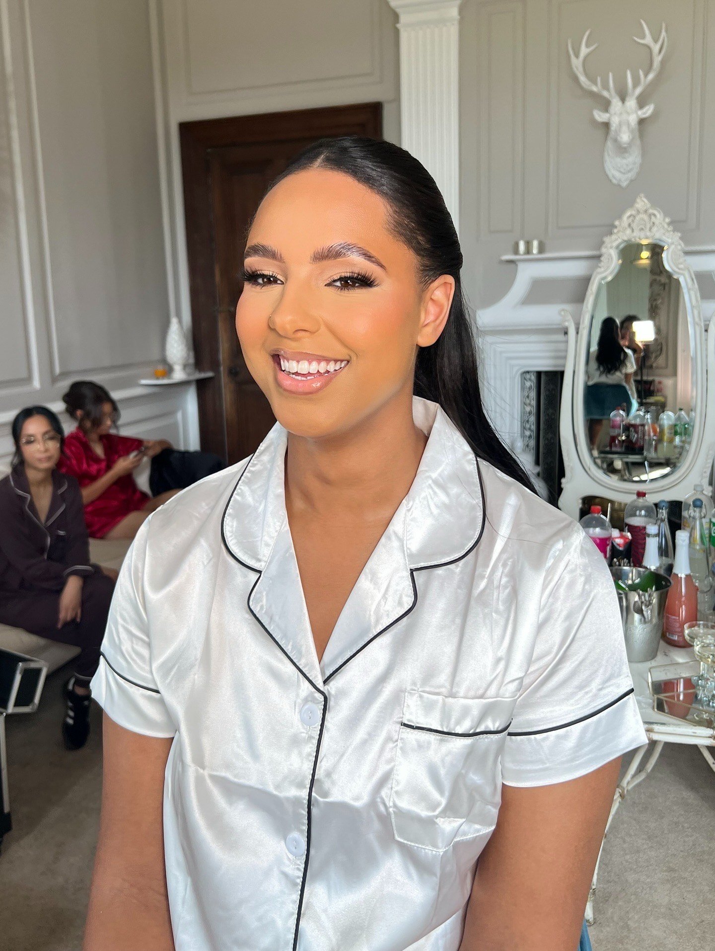 A woman smiling, wearing white satin pajamas with black piping, getting ready in a luxurious room with ornate decor, including a white mounted deer head, a mirror, and bottles of drinks on a counter.