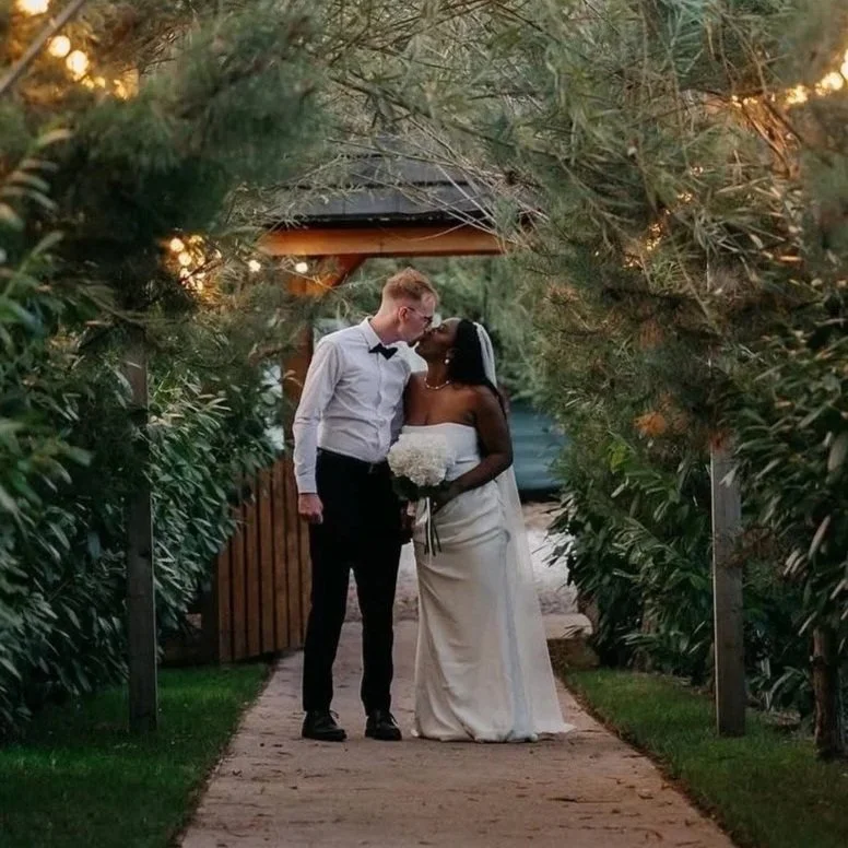 A newlywed couple stands on a garden path, kissing under a canopy of trees and greenery, with a small wooden structure in the background.