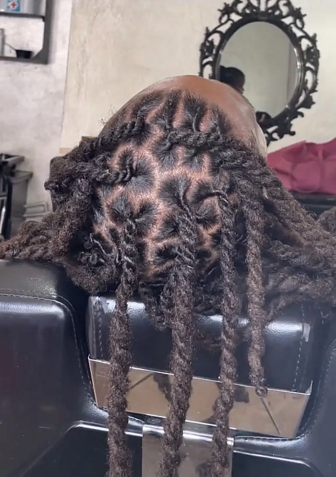 Close-up photo of a person with long, thick, dreadlocked hair hanging over a black chair or table, with a mirror and wall in the background.