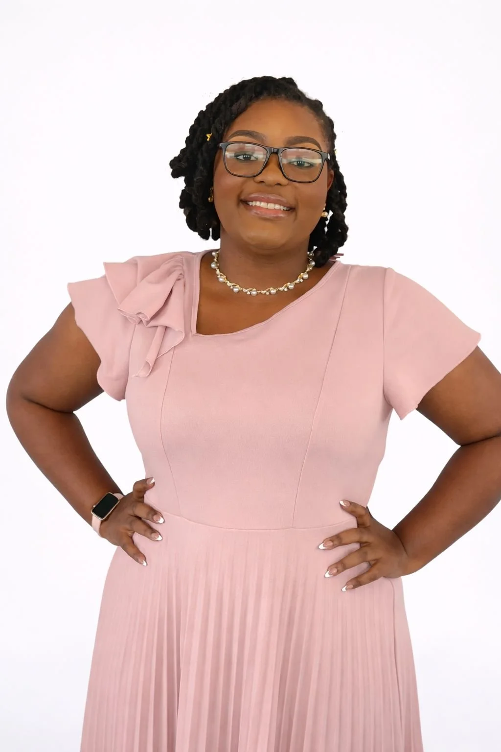 A woman wearing glasses and a pink dress, smiling with hands on hips against a white background.