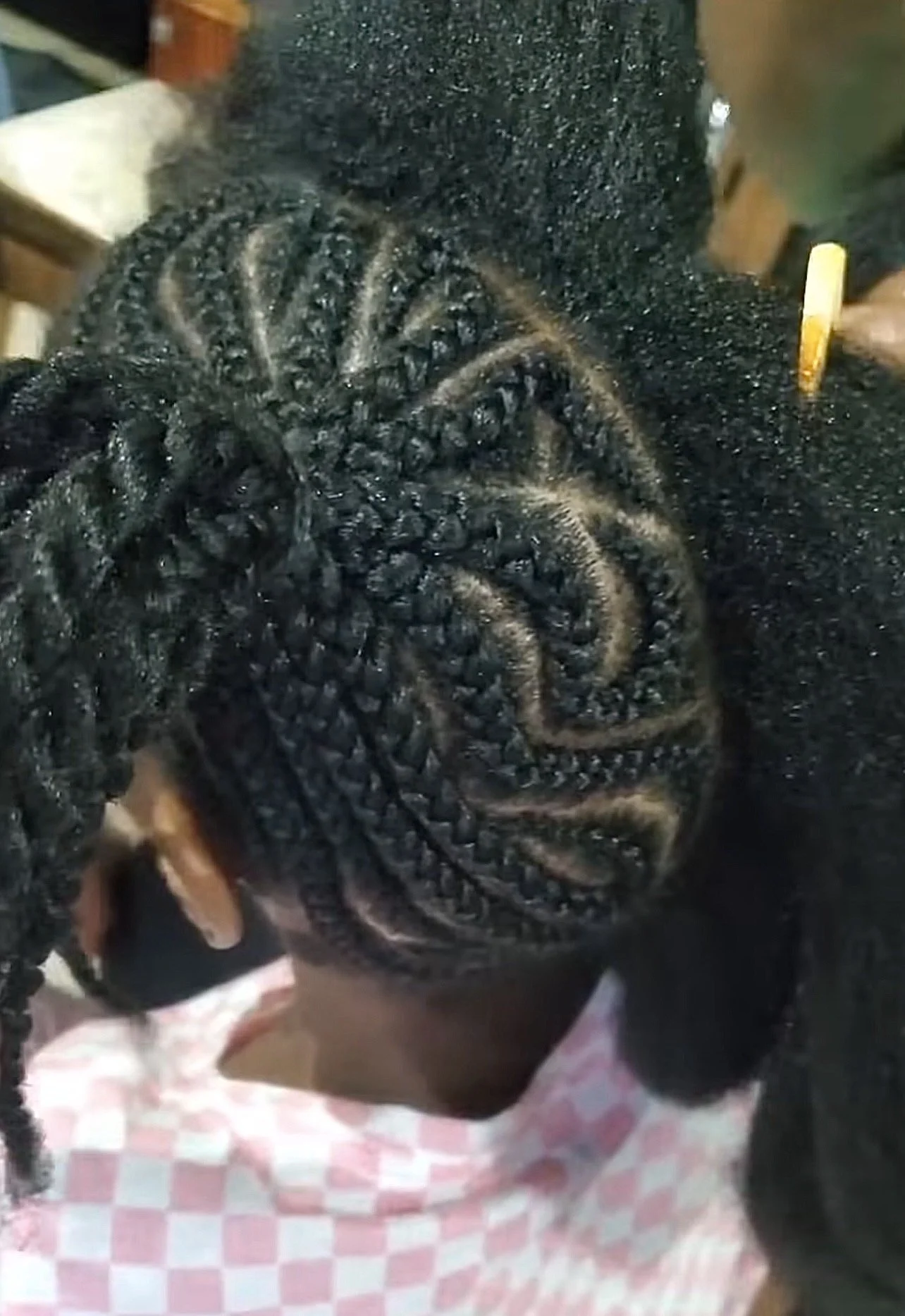 Close-up of a person's head with freshly braided hair, showcasing intricate cornrows and a natural black hairstyle, sitting on a pink and white checkered surface.