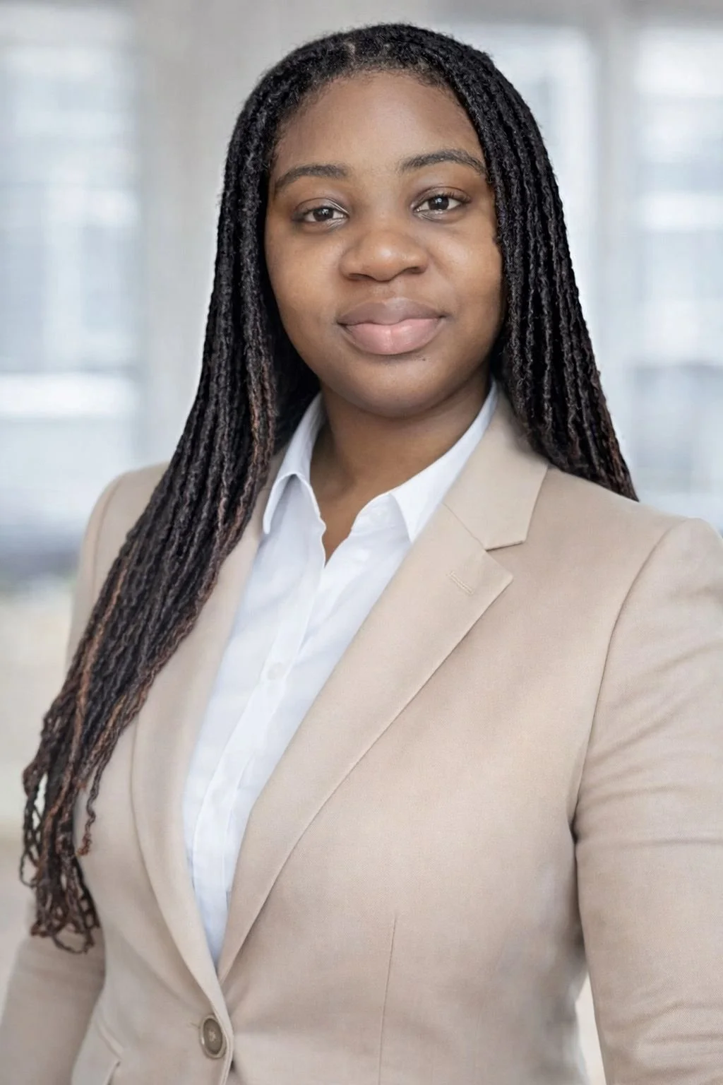 Professional woman with long braided hair wearing a beige blazer and white shirt, standing in a bright office environment.