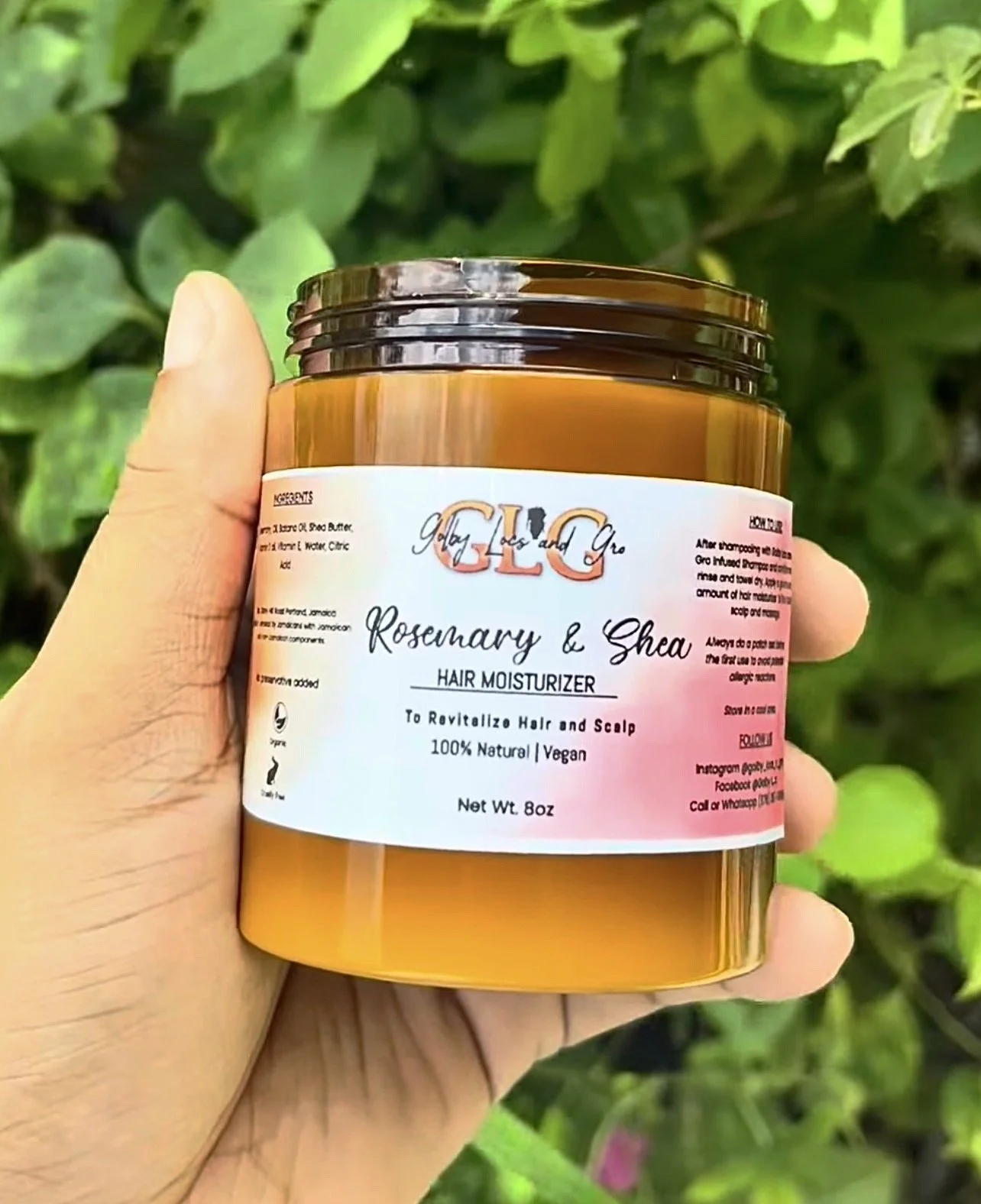A person holding a brown jar of herbal hair moisturizer labeled "Glo" with ingredients including rosemary and shea. The background features green foliage.