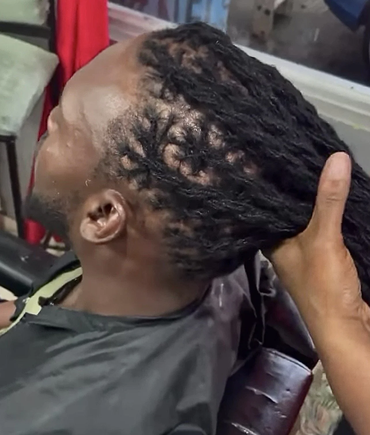 A person with long twisted dreadlocks sitting in a barber's chair, getting their hair styled or retwisted while a hand holds the hair section.
