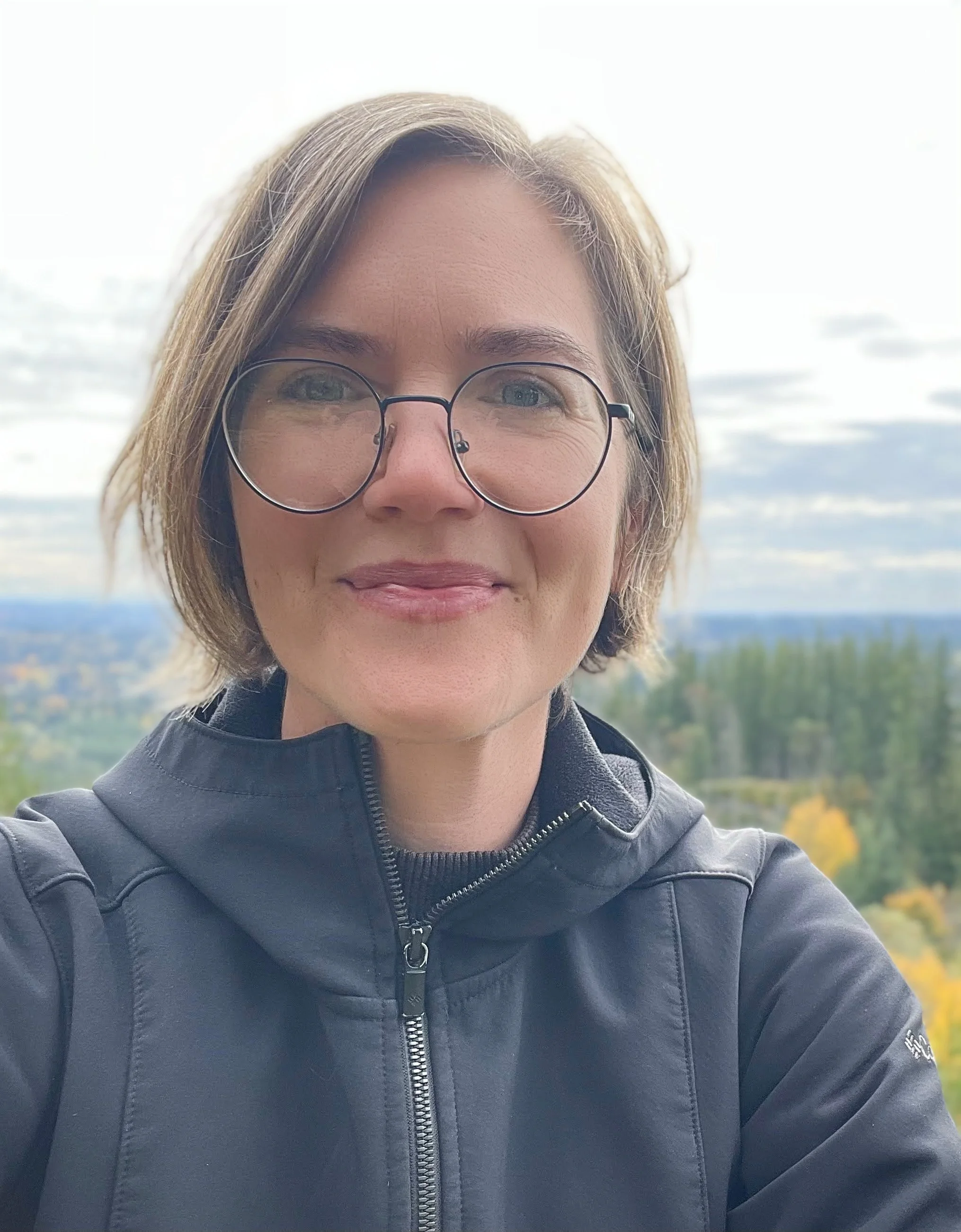 Jamie Clayton - A woman with short brown hair and glasses smiling outdoors with a scenic landscape of trees and sky in the background.