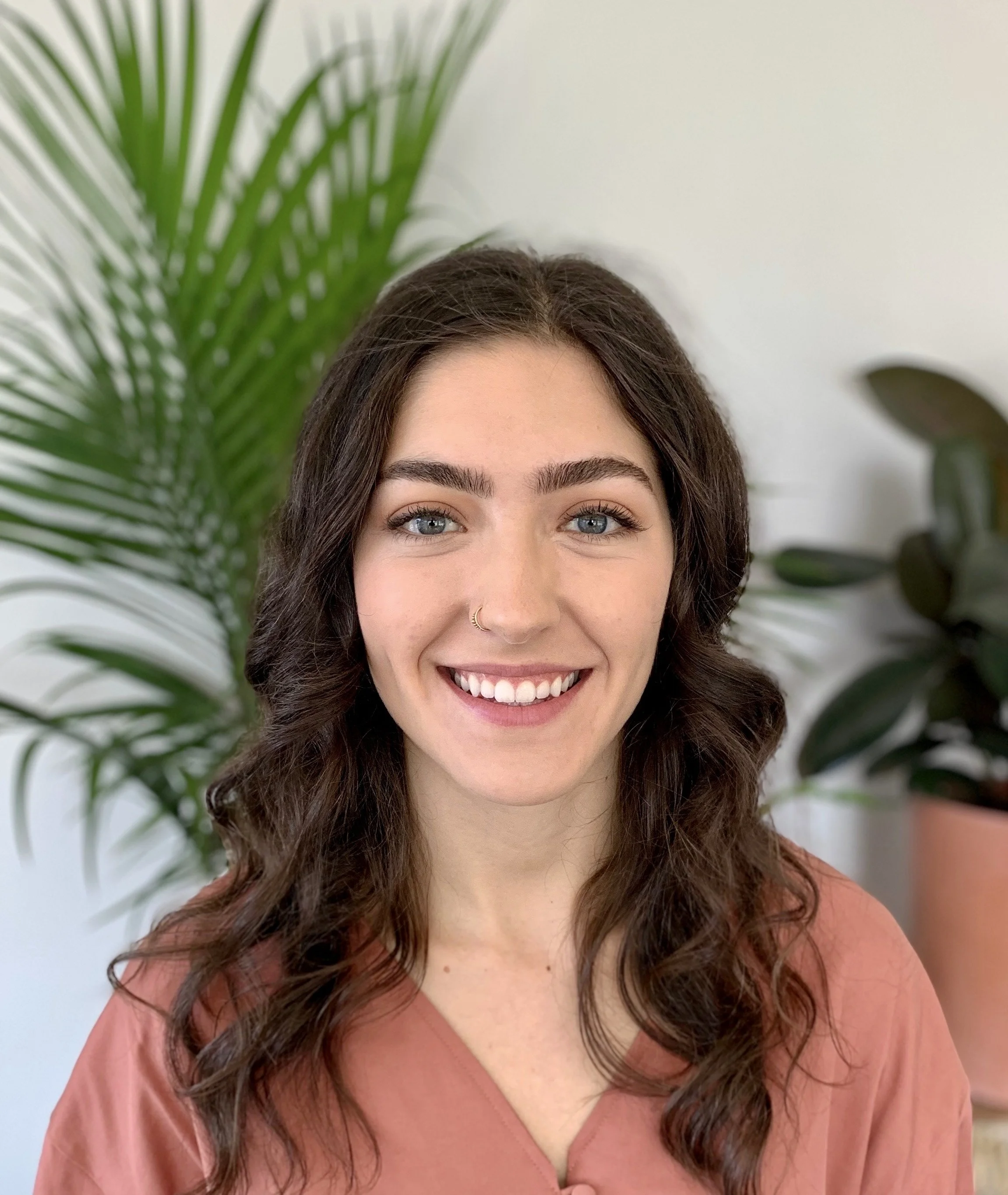 Maria Taylor - A woman with wavy brown hair, blue eyes, and light skin, smiling indoors with green plants in the background.
