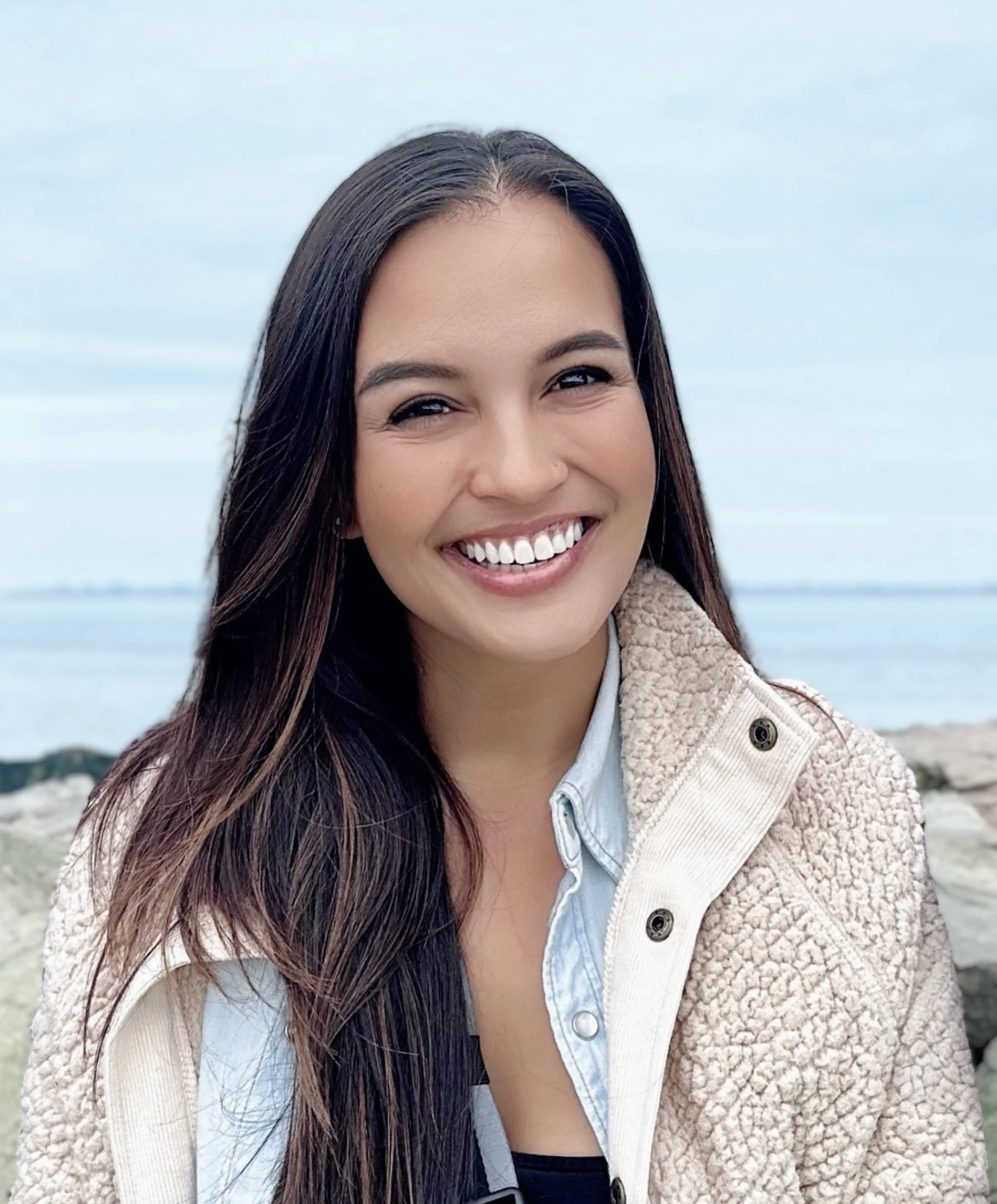 Rochelle Adriano - A woman with long dark hair, smiling, outdoors near a body of water and rocks, wearing a beige fleece jacket over a blue collared shirt.
