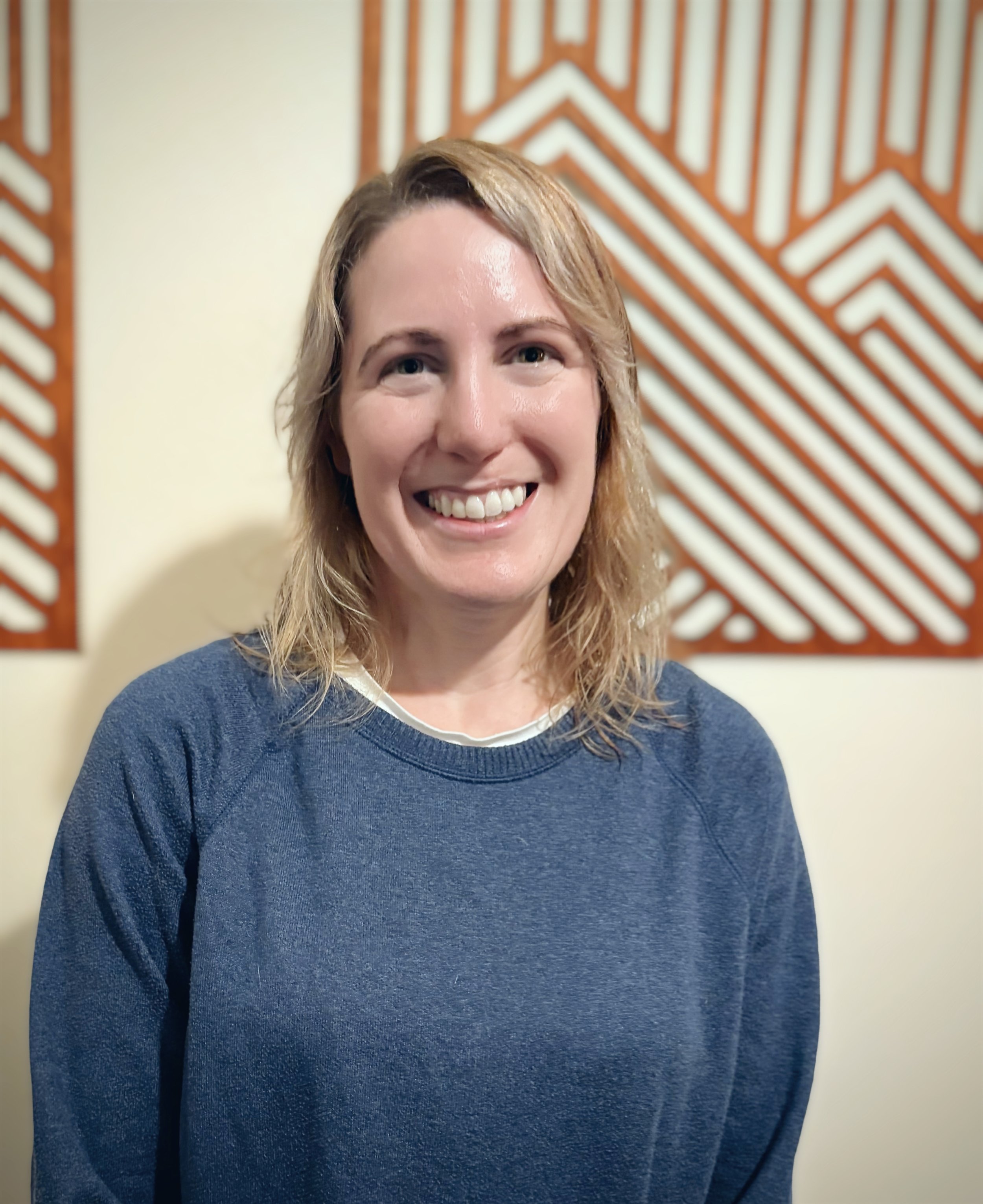 Geanna Revell - A woman with shoulder-length blonde hair, smiling, wearing a blue sweater, standing indoors with patterned wall art behind her.