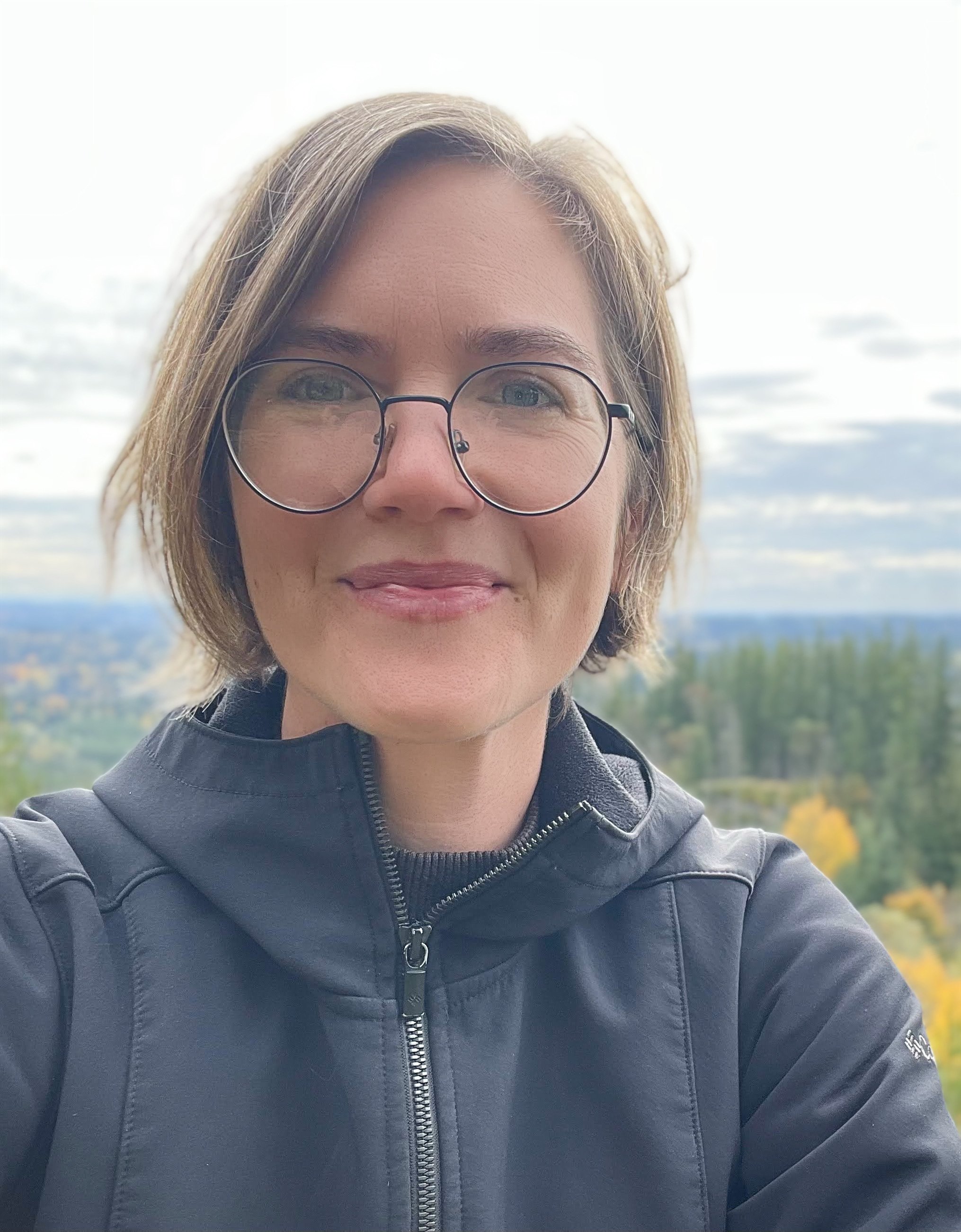 Jamie Clayton - Close-up of a woman with short brown hair and glasses, smiling outdoors with a scenic landscape of trees and sky in the background.