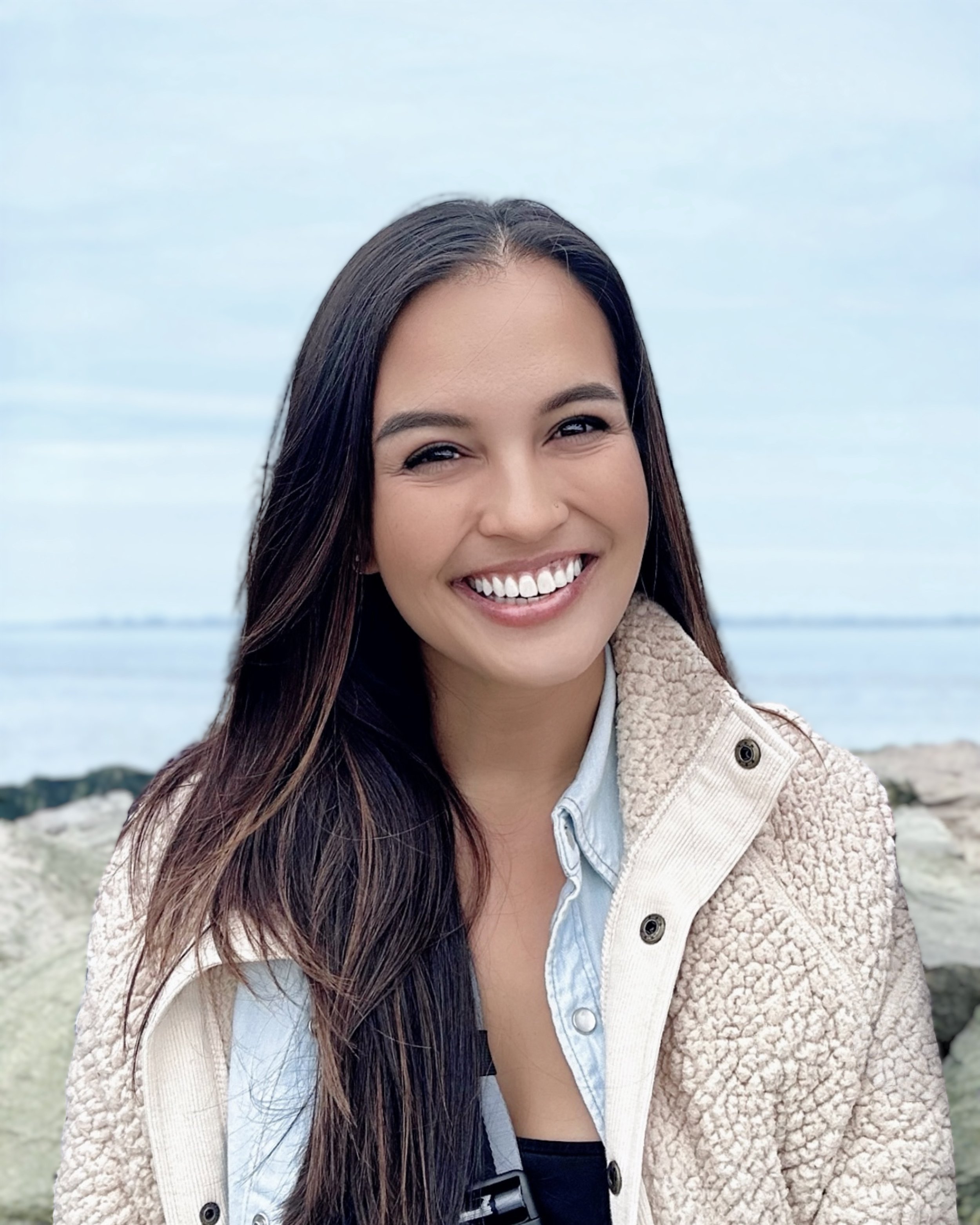 Rochelle Adriano - A smiling woman with long dark hair standing outdoors near water and rocks.