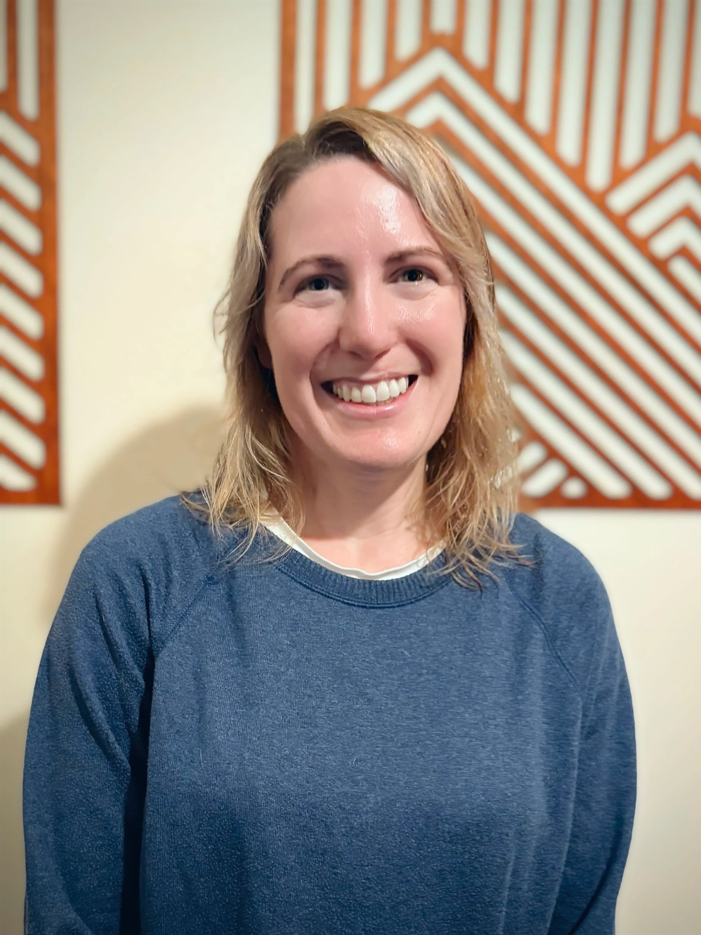 Geanna Revell - A smiling woman with shoulder-length blonde hair wearing a blue sweatshirt standing in front of a wall with decorative wooden panels.