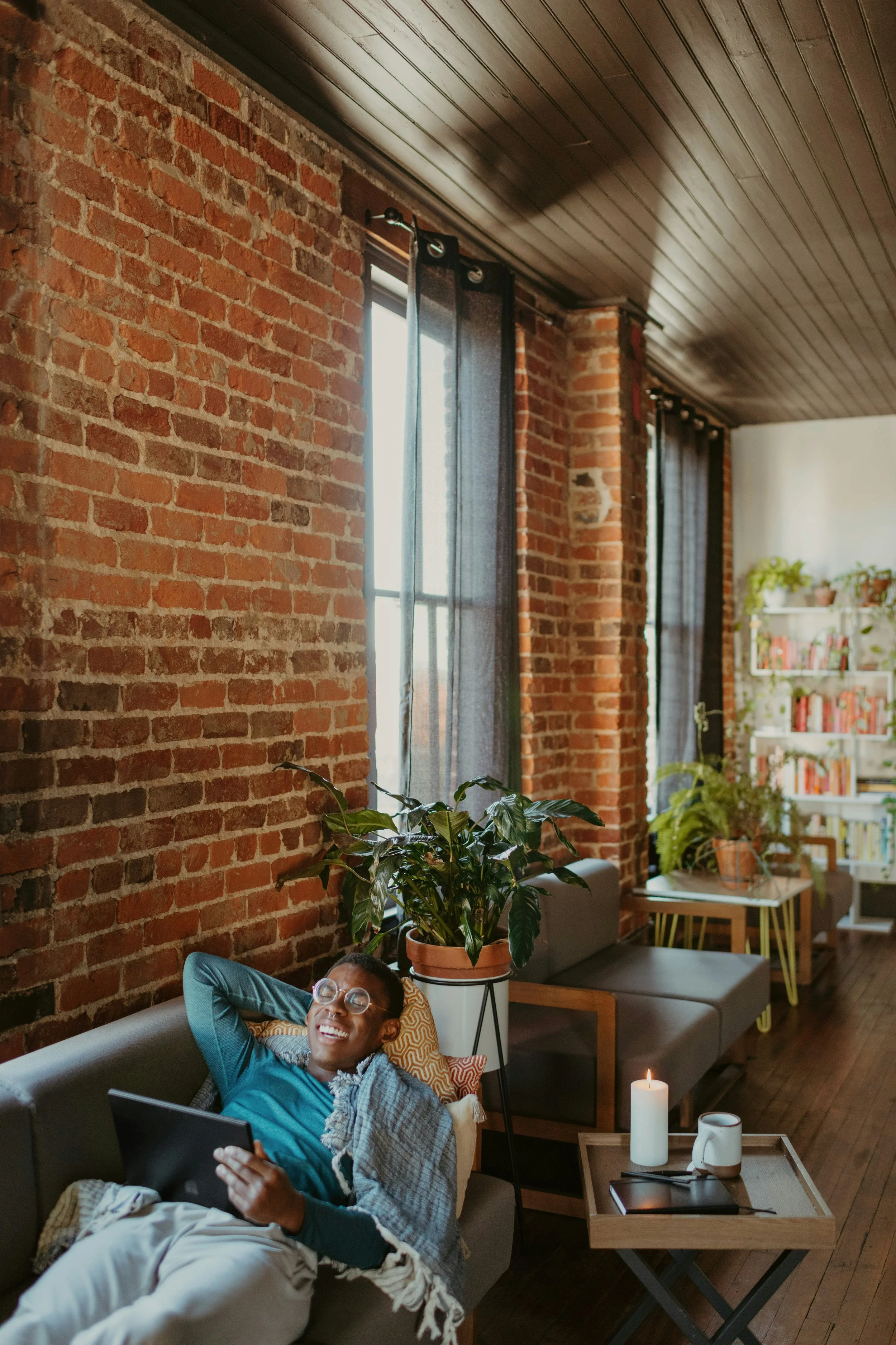 A person relaxing on a sofa in a cozy living room with brick walls, large windows with black curtains, plants, bookshelf, and a small tray table with a candle and mug.