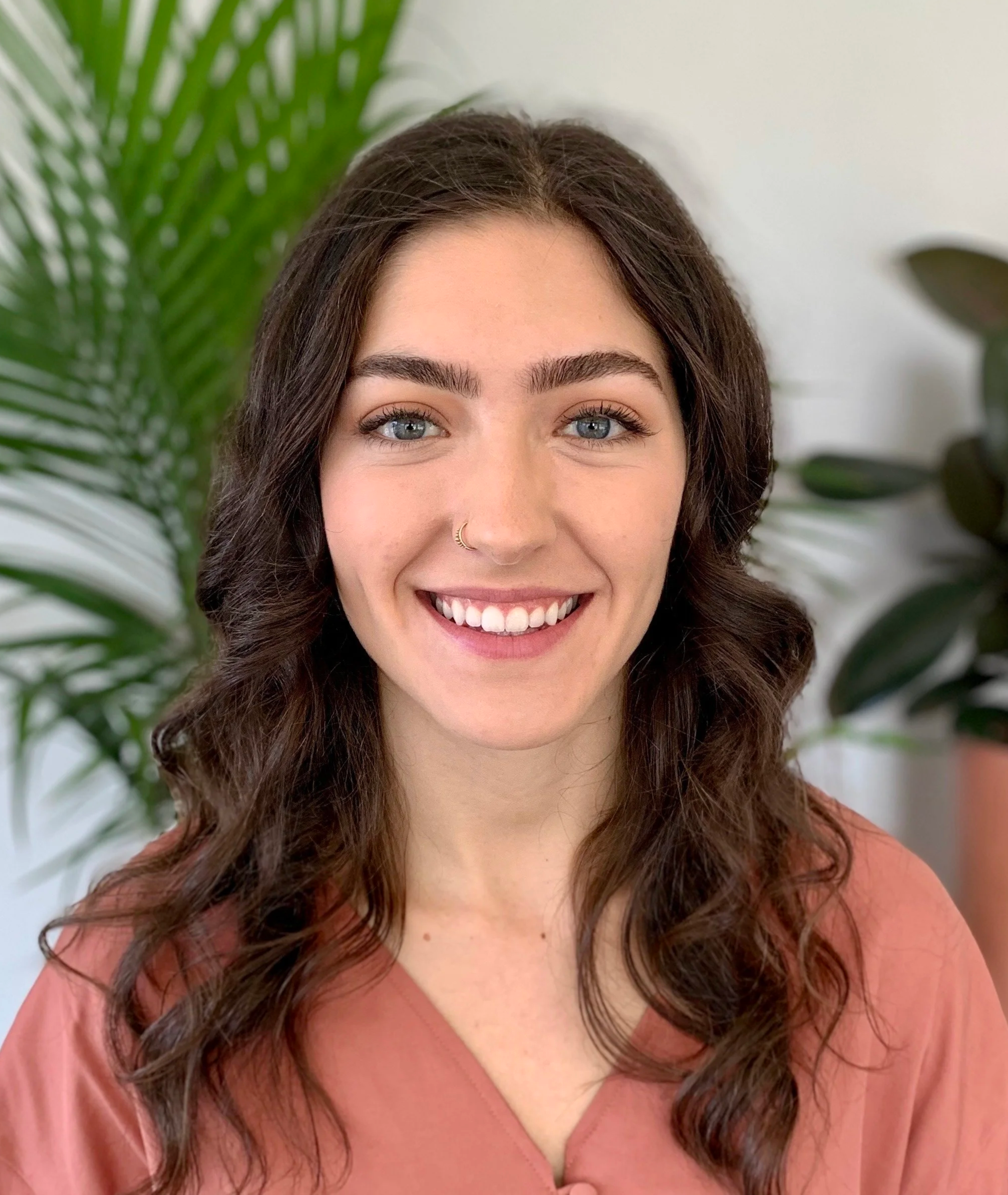 Maria Taylor - A woman with wavy brown hair, blue eyes, and a nose ring smiling in front of green plants.