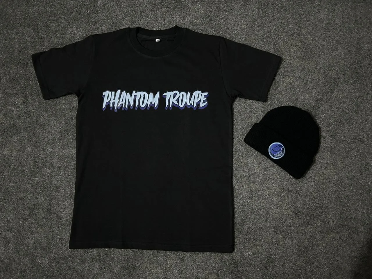 Black T-shirt with 'PHANTOM TROUPE' written in stylized blue and white letters, and a black knit beanie hat with a space-themed logo on a gray textured surface.
