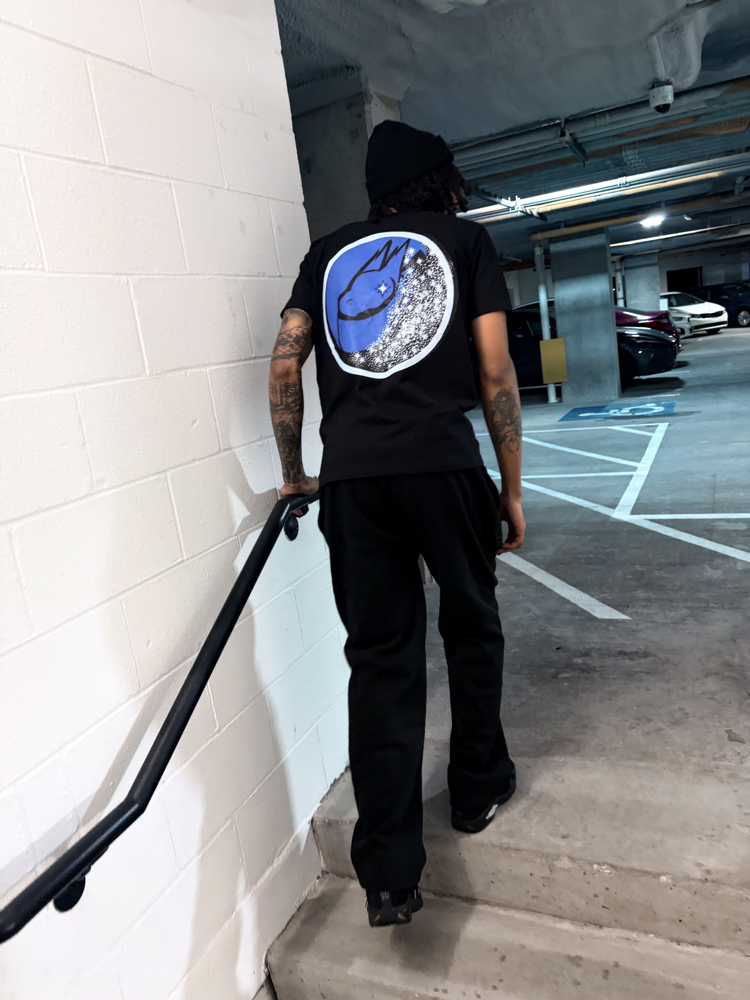 A person with tattoos wearing a black beanie, black t-shirt with a blue and white graphic on the back, black pants, and shoes, walking down a stairway in a parking garage.
