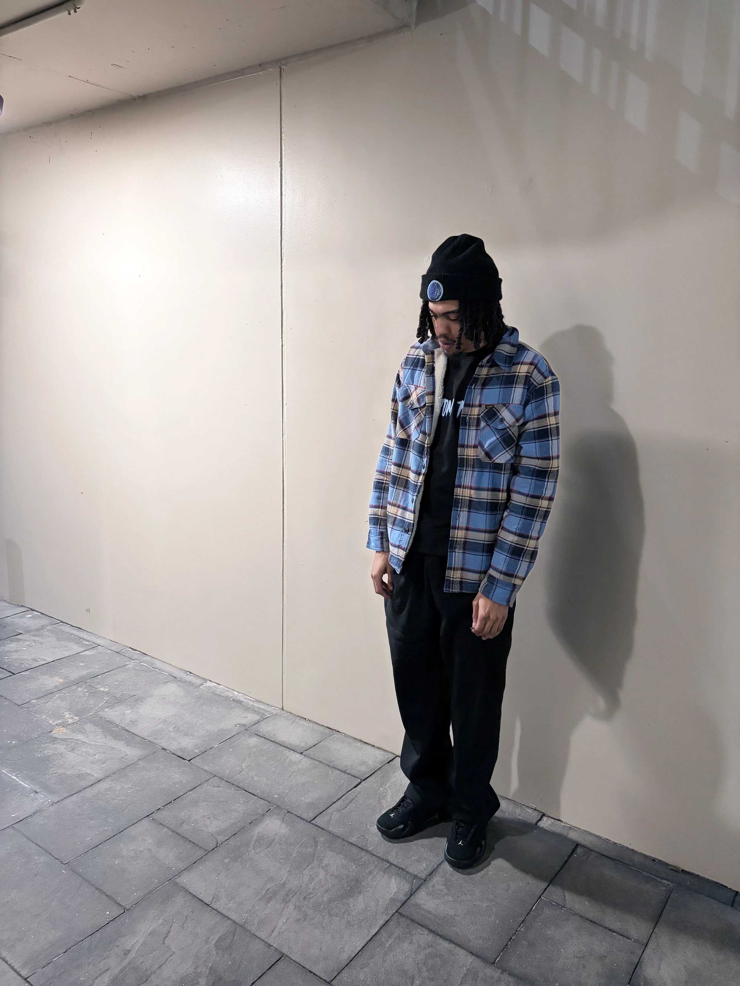 A person standing against a beige wall, wearing a black beanie, glasses, a plaid shirt over a black T-shirt, black pants, and black shoes, looking down with hands loosely hanging by their sides.