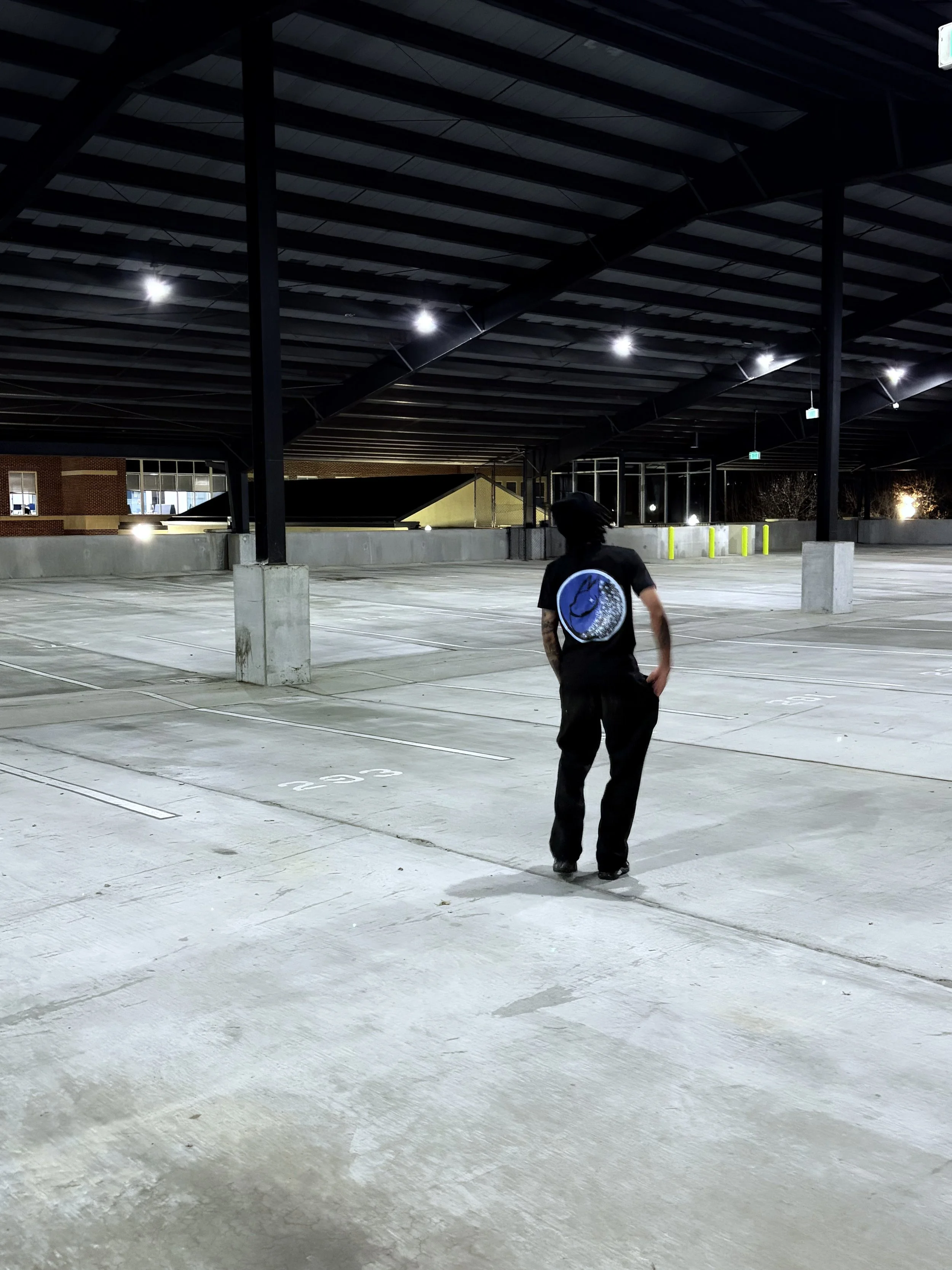 A person walking alone in an empty, well-lit parking garage at night, wearing a black t-shirt with a blue and white logo on the back.