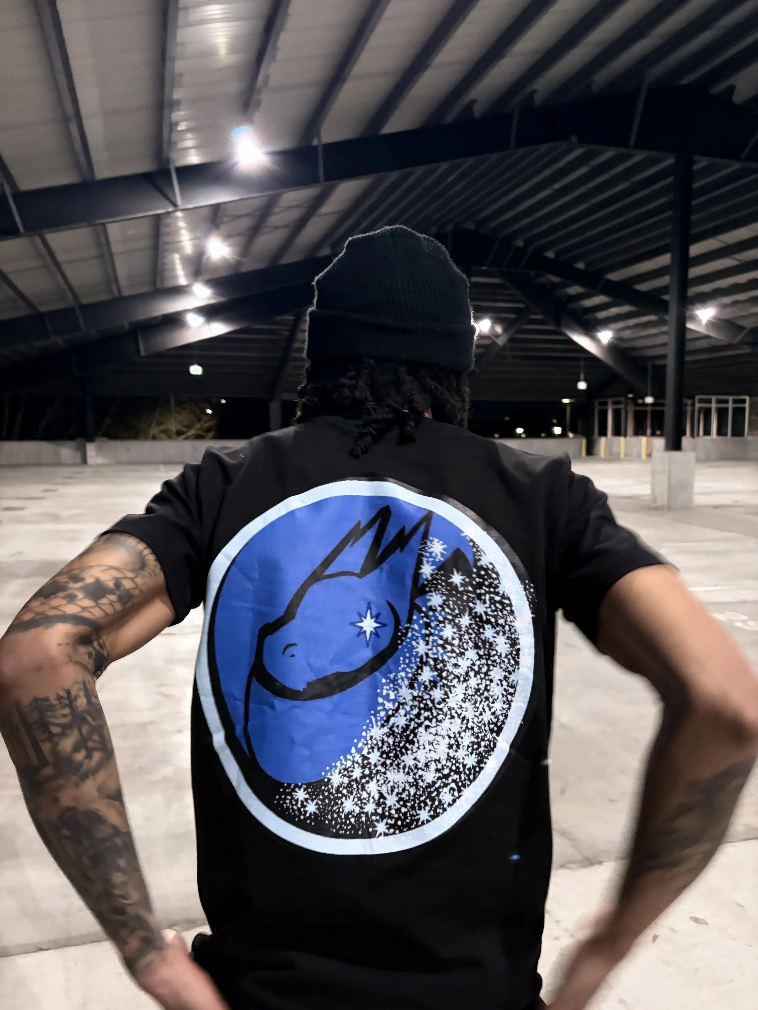 Person wearing a black beanie and black t-shirt with a large blue and white graphic on the back, standing in an empty parking garage at night.