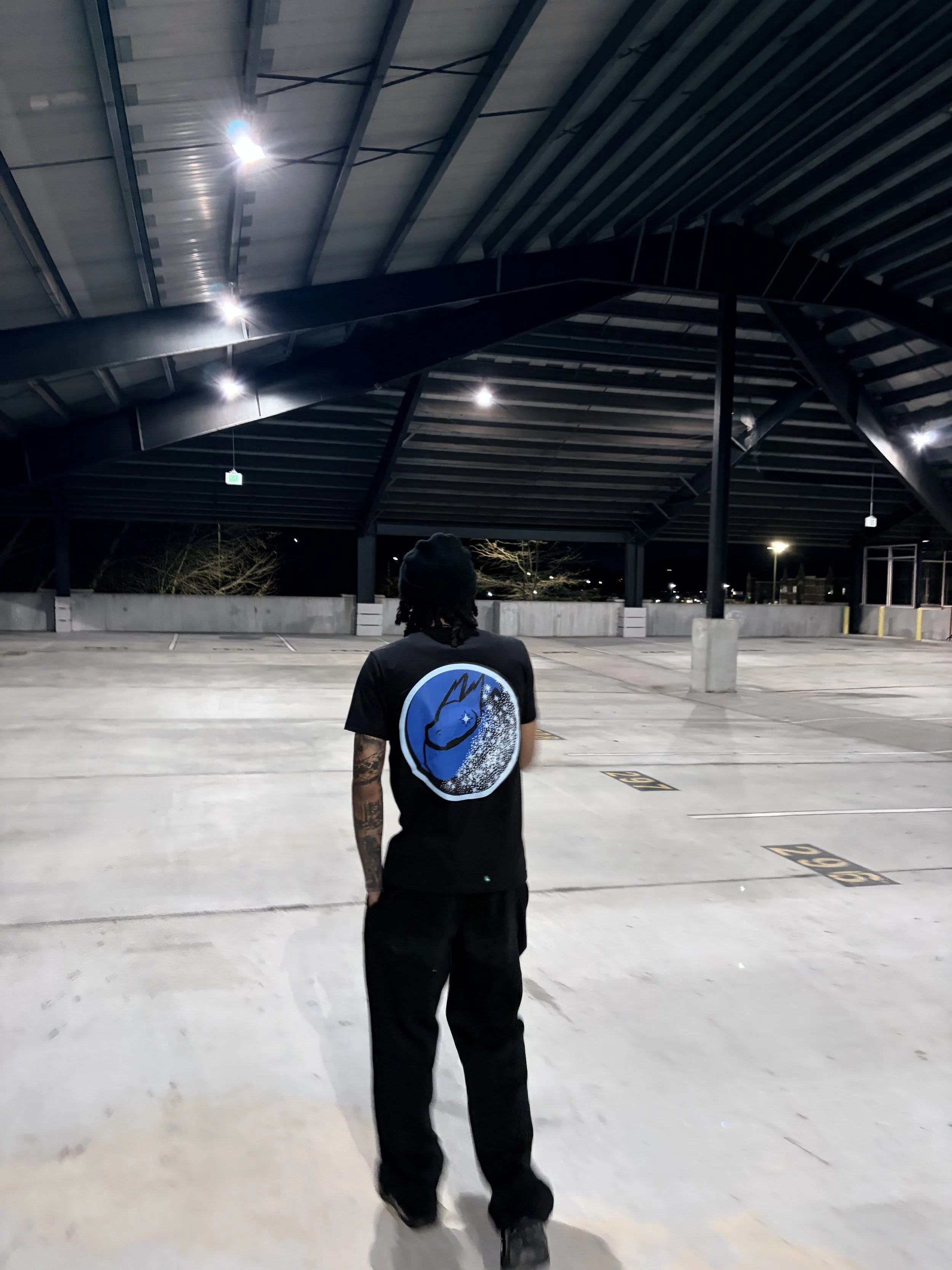 A person standing alone in an empty, well-lit parking garage at night, wearing a black t-shirt with a blue graphic on the back and black pants.