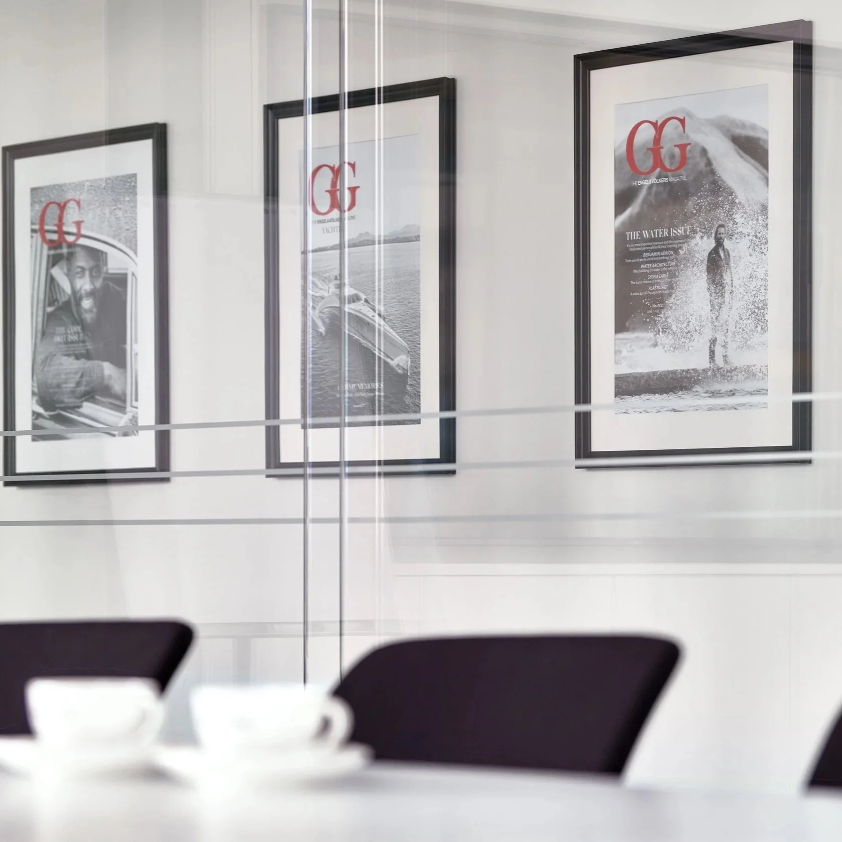 Framed magazine covers on a wall viewed through a glass partition, with part of a table and cups in the foreground.