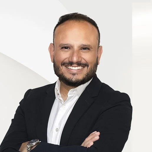 A smiling man with a beard, wearing a white shirt and a black blazer, standing against a white background.