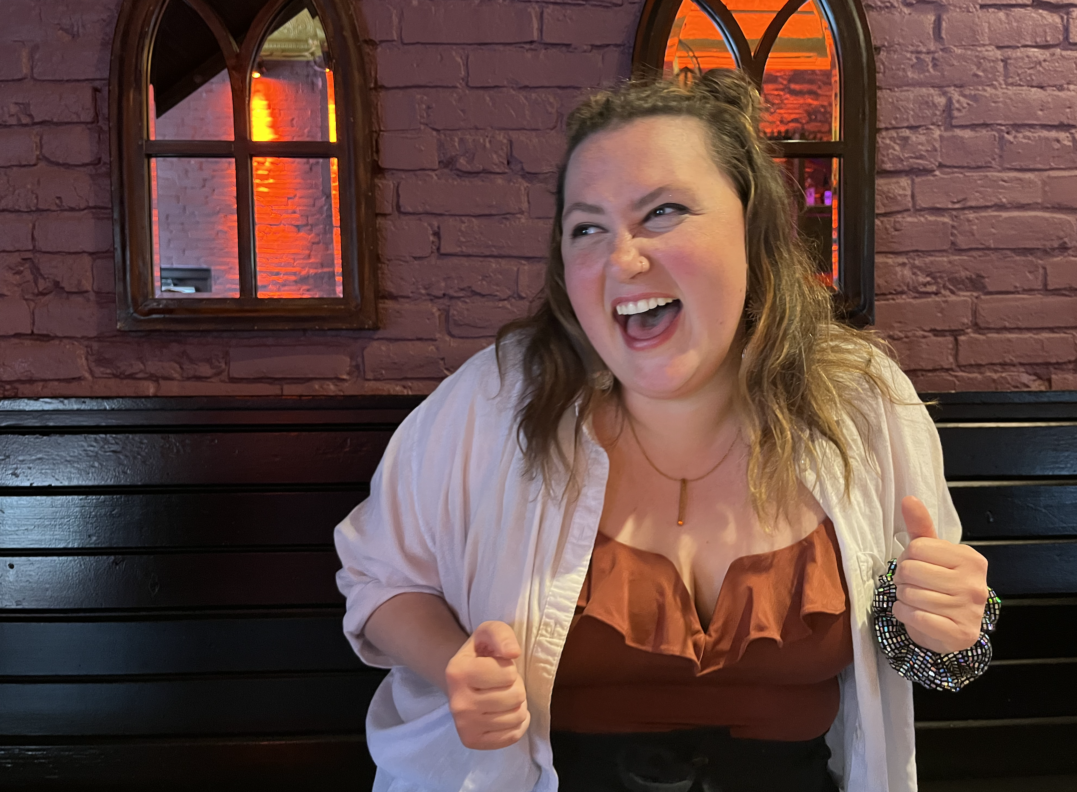 A woman with wavy brown hair, smiling and making a fist gesture, seated in a restaurant with purple brick walls and framed arched mirrors with orange and purple lighting.