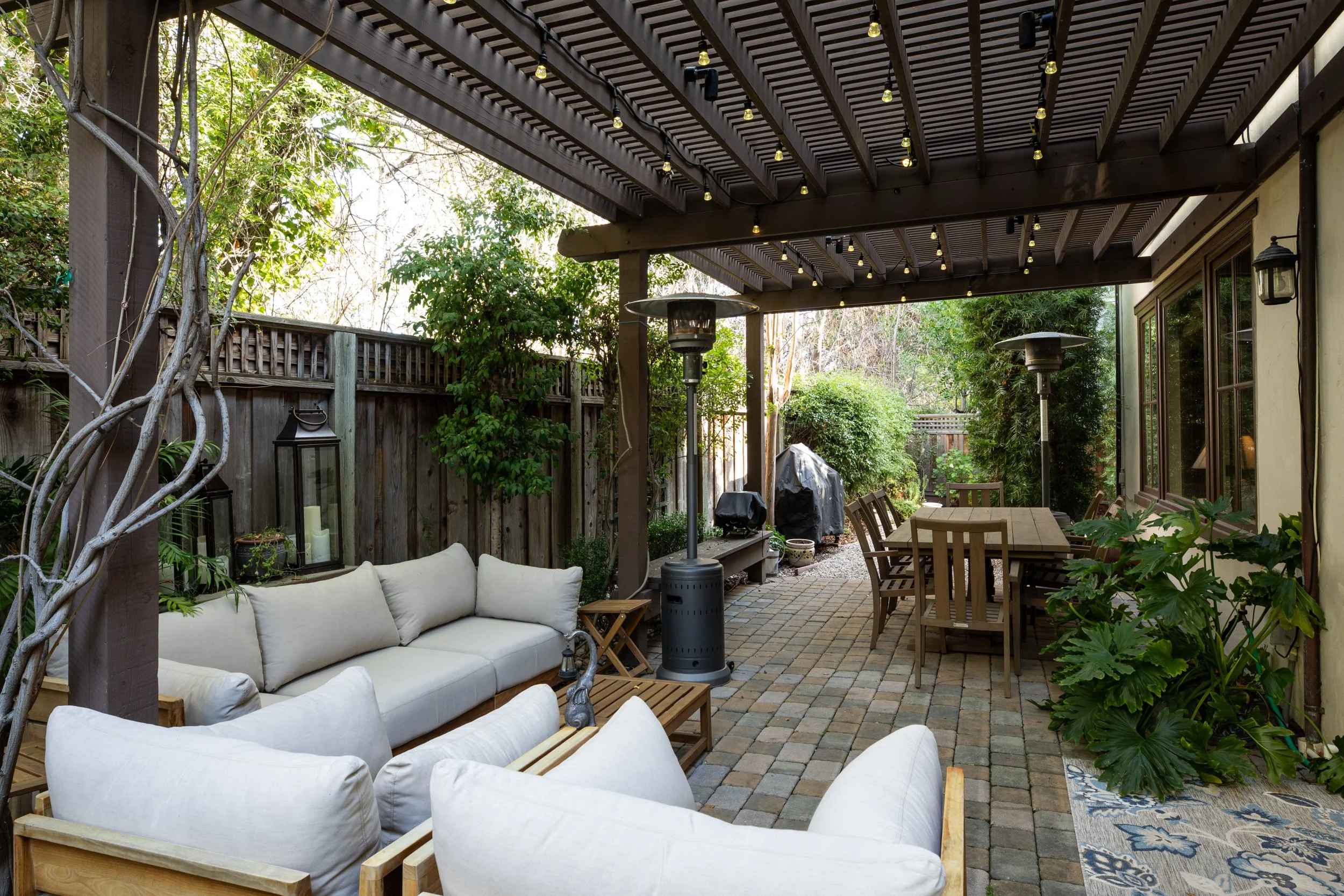 Backyard patio with outdoor furniture, table, stove, and surrounded by greenery and a wooden fence.