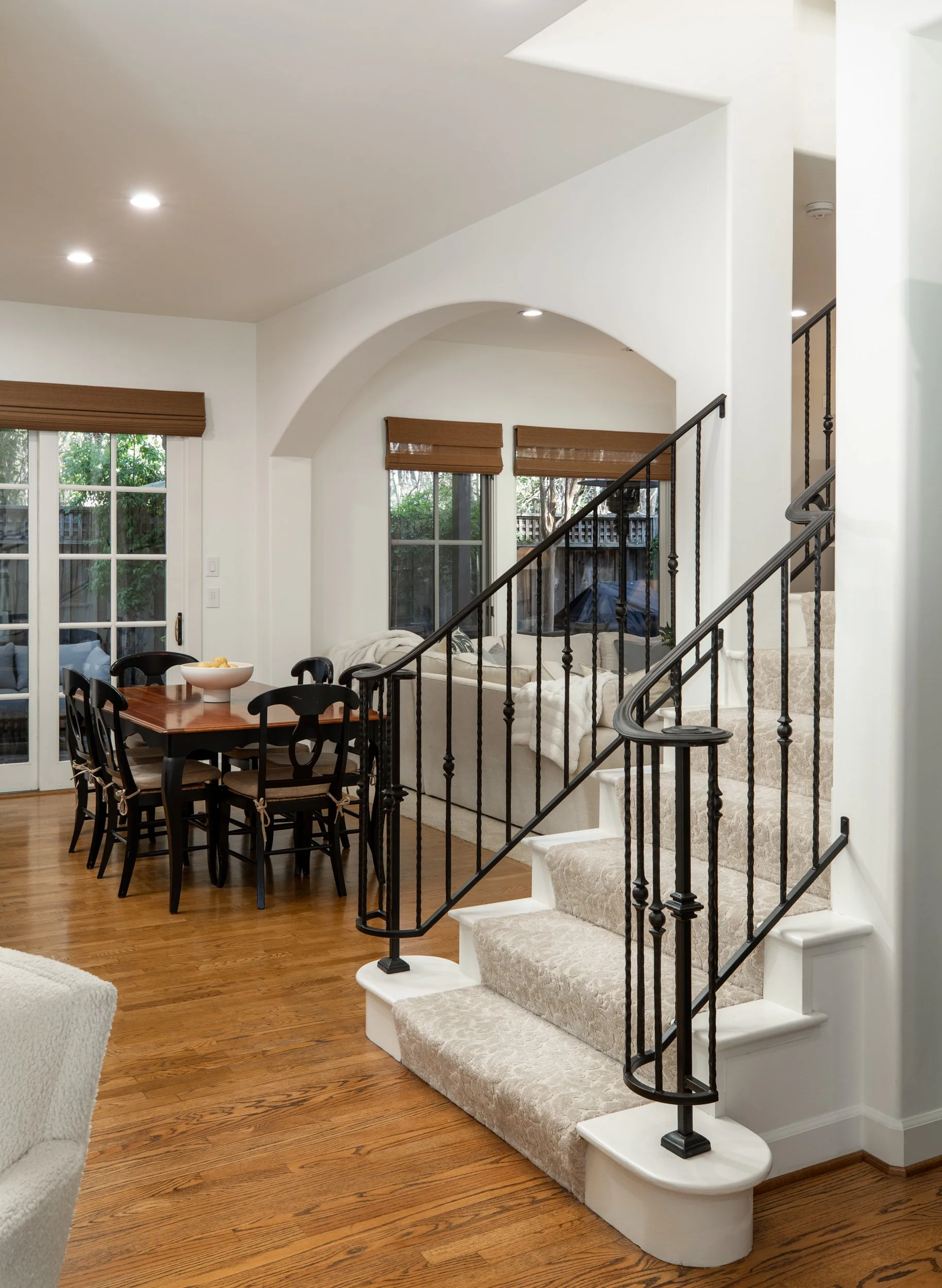 Interior of a house showing a staircase with black iron railings, a dining area with a wooden table and black chairs, large windows with brown blinds, and hardwood flooring.