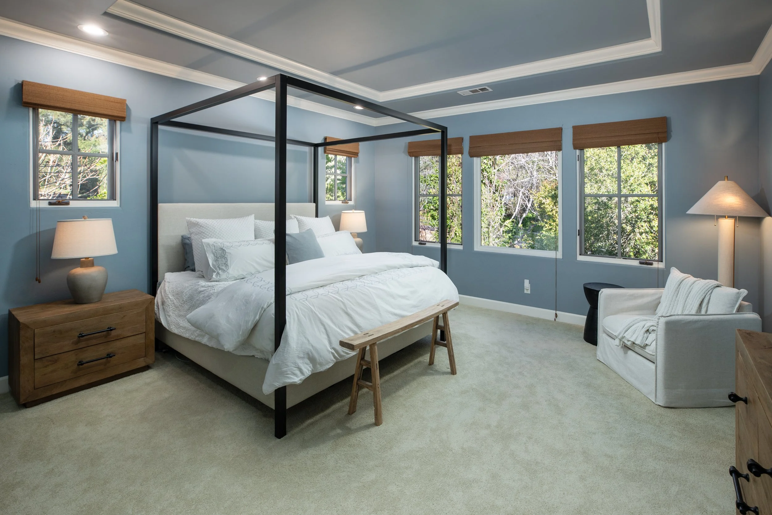 Bedroom with light blue walls, a white bed with a black canopy frame, wooden nightstands with lamps, a white armchair with a blanket, and multiple windows showing trees outside.