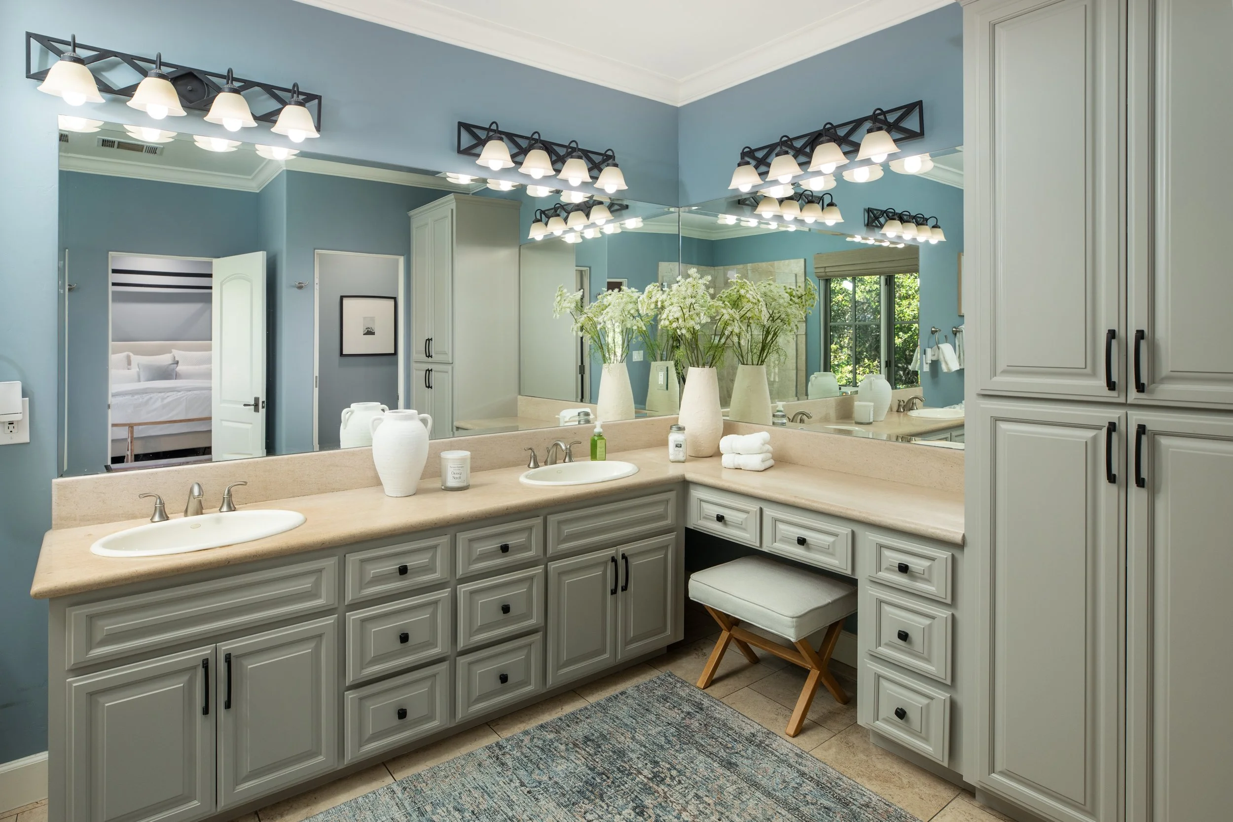Bathroom with dual sinks, large mirror, white cabinetry, counters with vases, towels, and toiletries, and a view into a bedroom through an open door