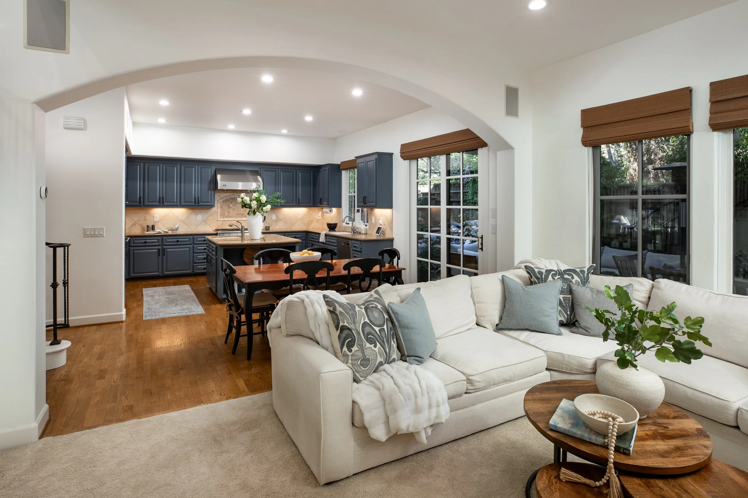 Open-concept living room and kitchen with white sectional sofa, wooden coffee table, and blue kitchen cabinets, large windows with brown blinds, hardwood floors, and ceiling lights.