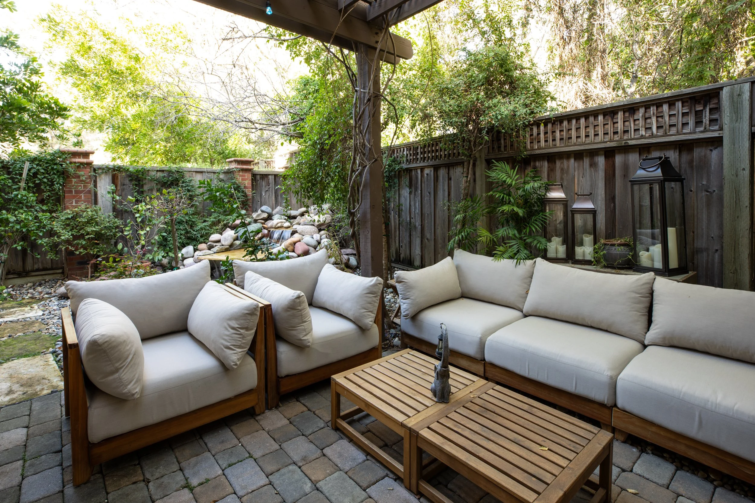 Outdoor patio with white cushioned seating, wooden coffee table, lanterns, greenery, and a small waterfall with rocks.