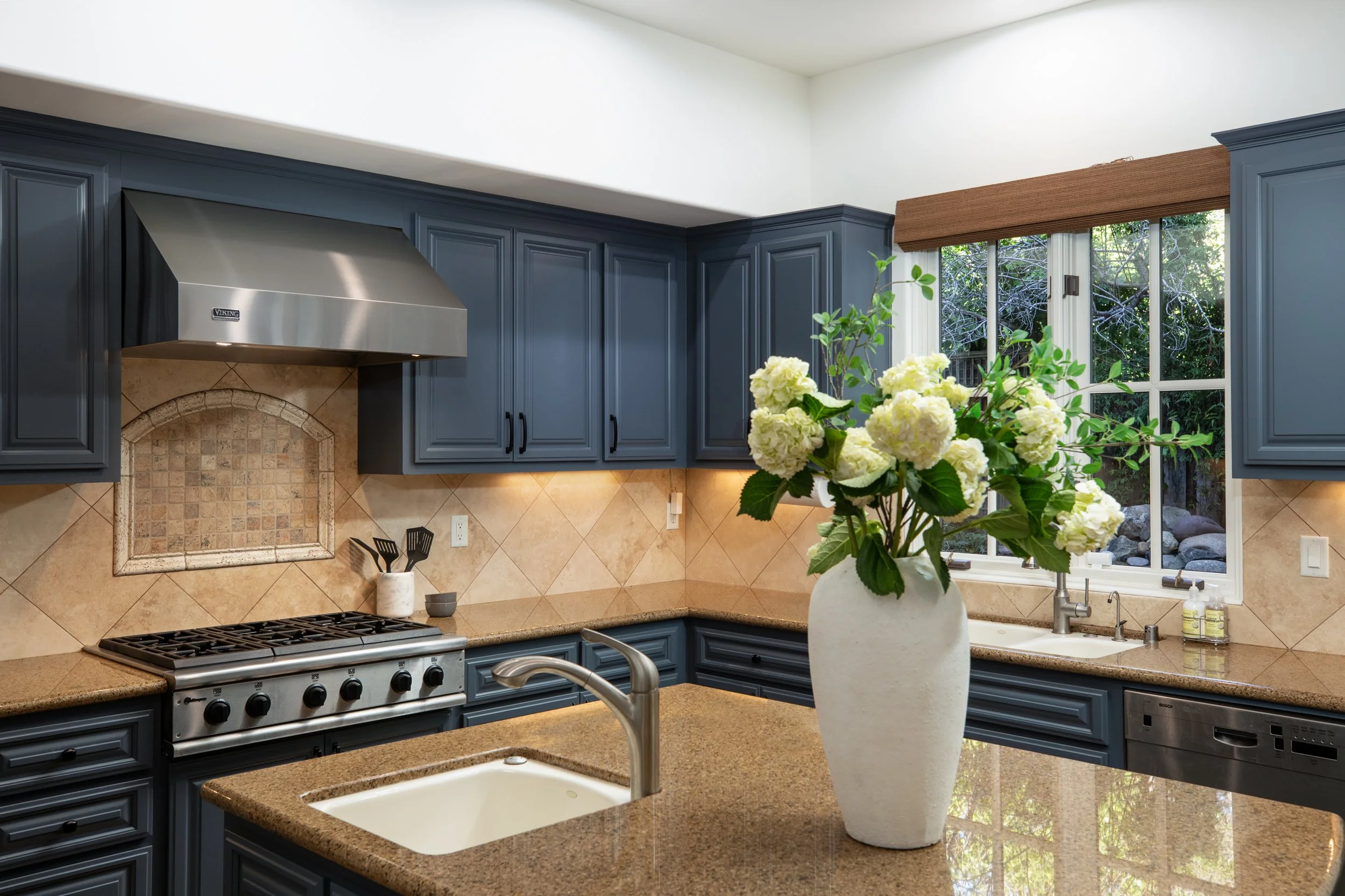 Modern kitchen with blue cabinets, a granite countertop, a vase of white hydrangeas, and a view of the outdoors through a window.