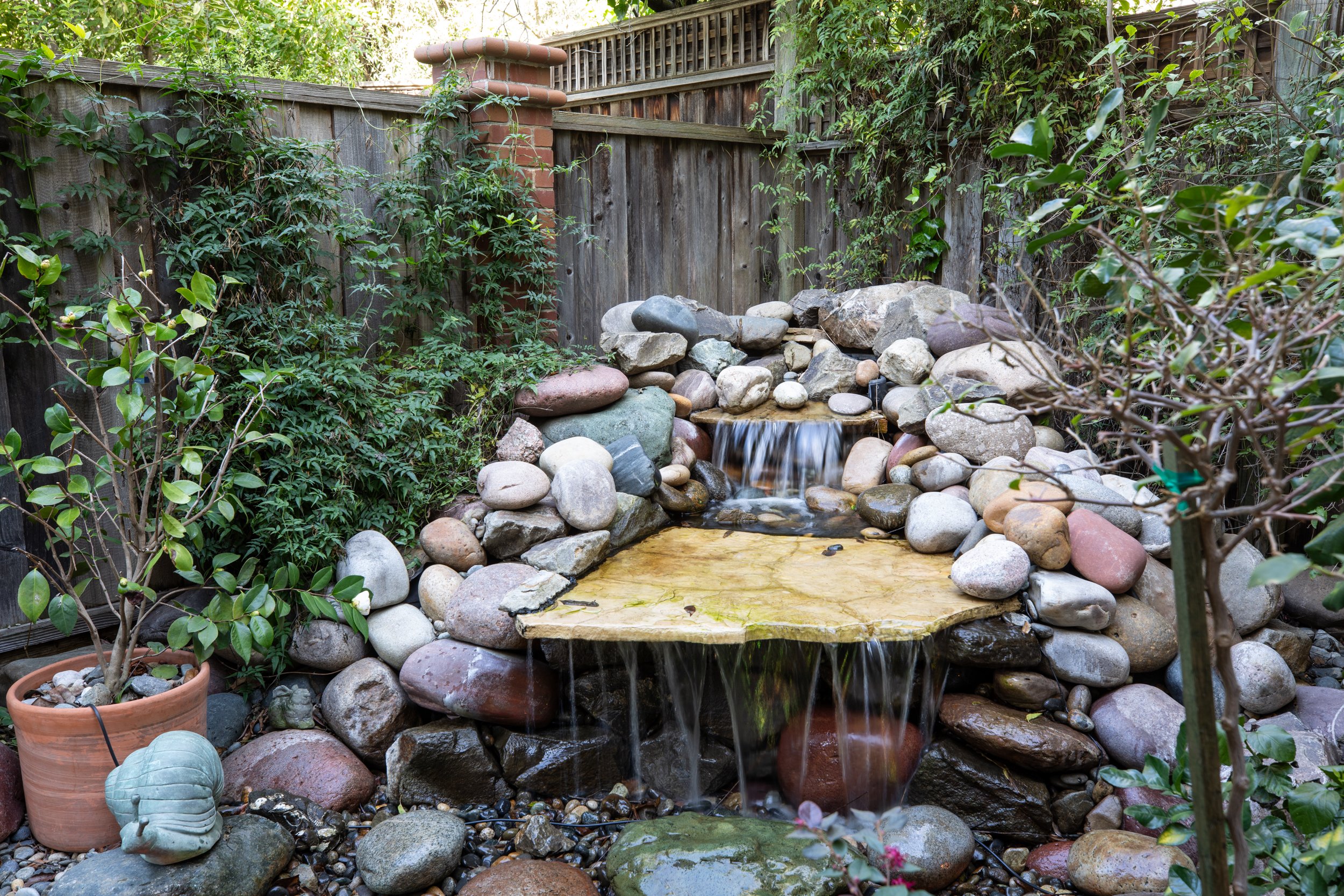 Small backyard pond with a waterfall, surrounded by rocks and plants, enclosed by a wooden fence.