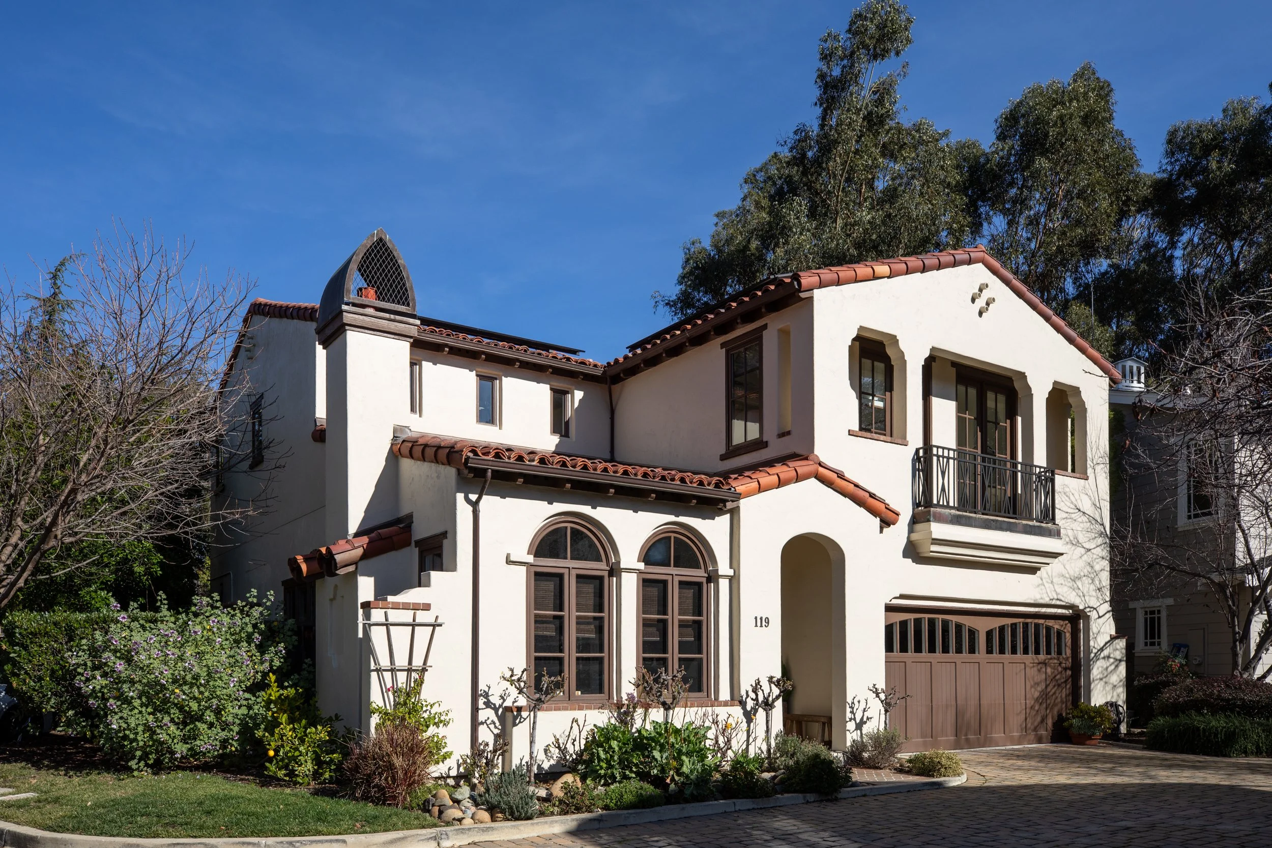 A white, two-story house with a red tile roof, arched windows, a small balcony, and a landscaped front yard with bushes and trees.