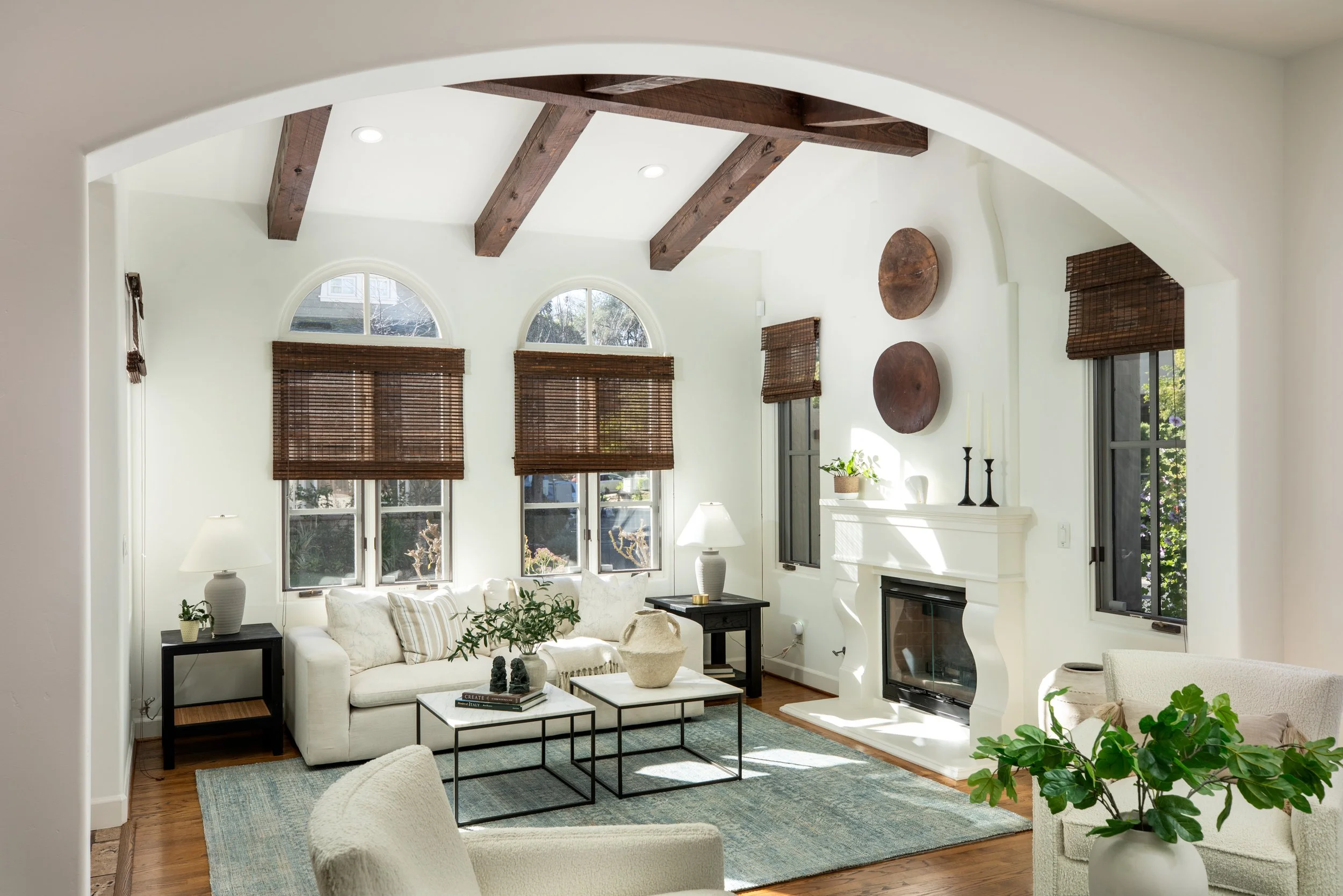 Bright living room with white walls, wooden ceiling beams, and hardwood floors, featuring a white sofa, black side tables, a fireplace with white mantel, and multiple windows with brown blinds.