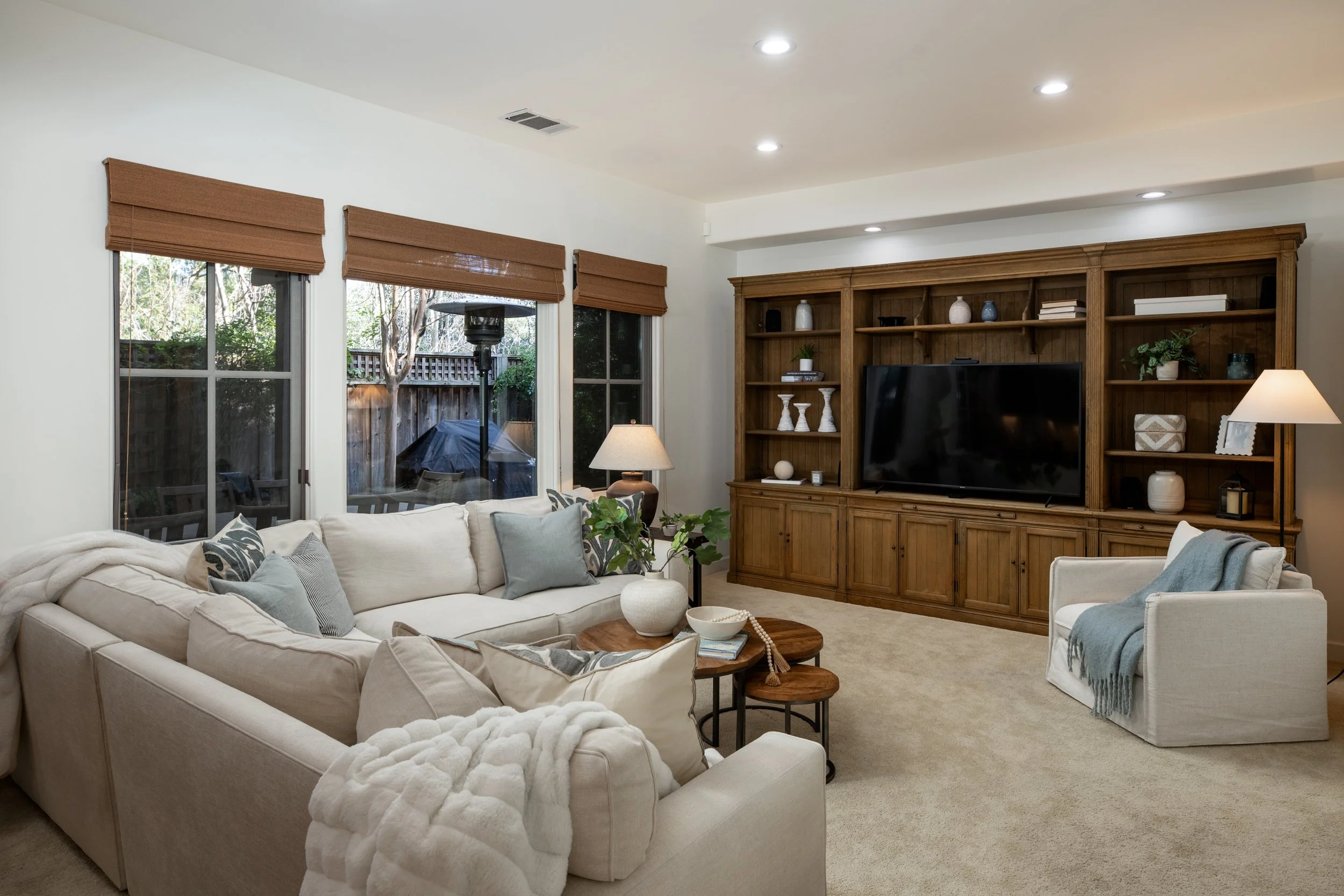 Living room with a large cream-colored sectional sofa, a wooden coffee table, and a white armchair. The room has large windows with brown shades, a beige carpet, and a dark wood entertainment center with a television, books, and decorative items. The