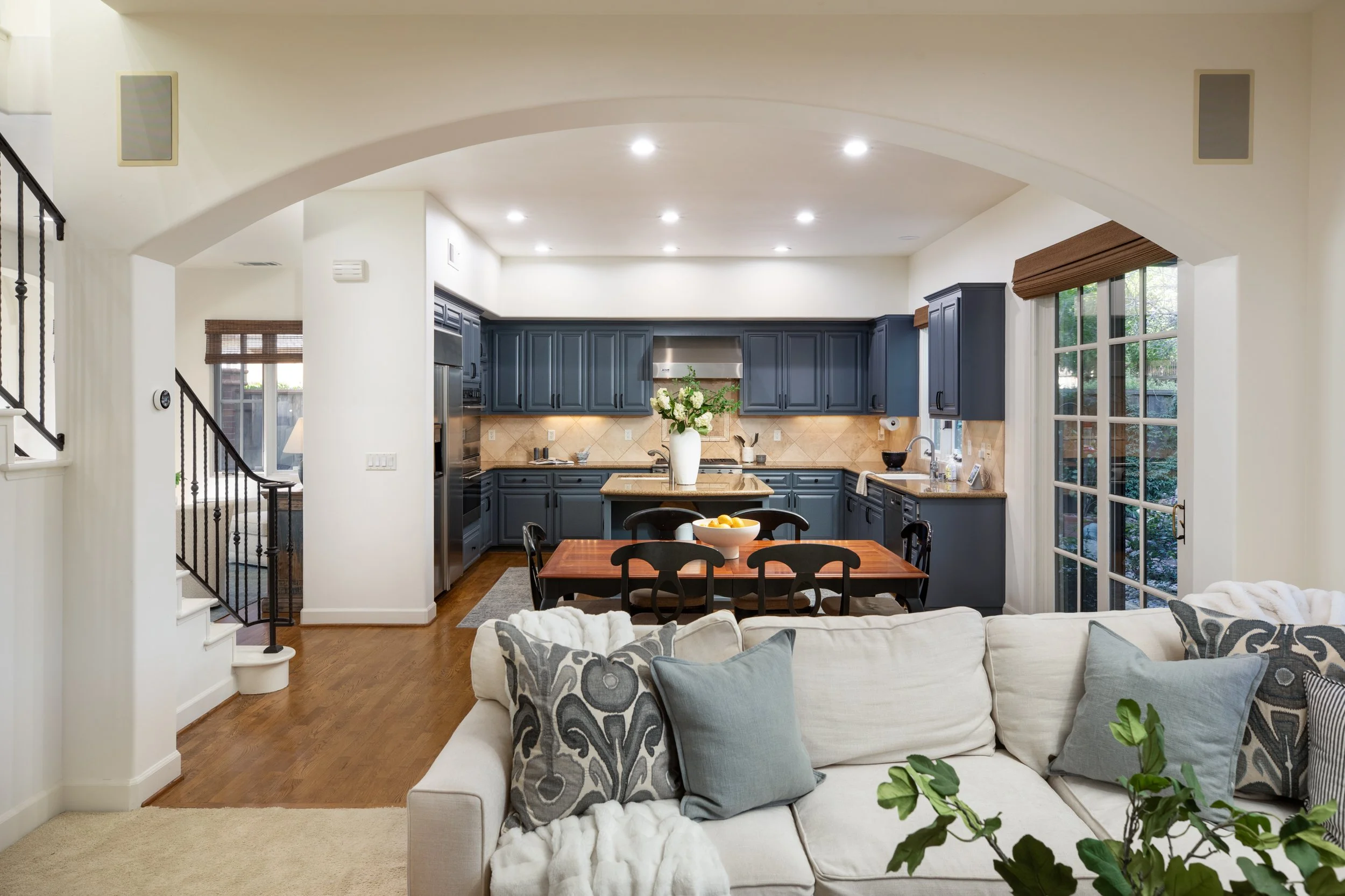 Open-concept living room and kitchen with navy blue cabinets, a wooden dining table with six black chairs, a white sofa with patterned and solid pillows, large windows with blinds, and hardwood flooring.