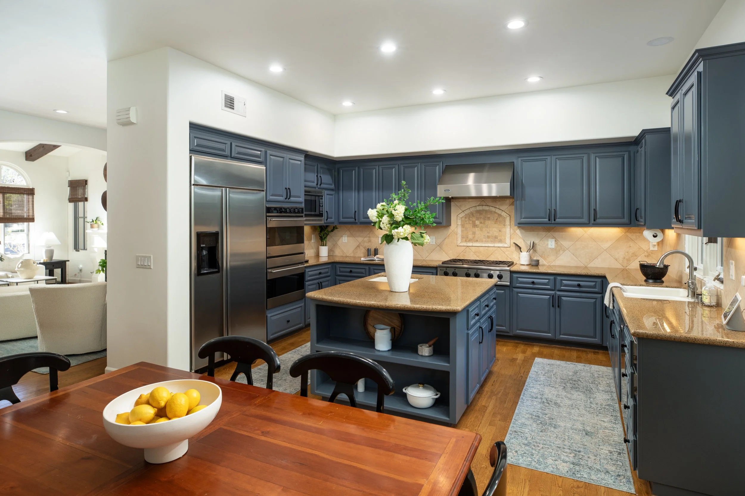 Modern kitchen with blue cabinets, beige countertops, stainless steel appliances including a refrigerator, oven, and microwave, and a kitchen island with a white vase of white flowers on top. There is a wooden dining table with a bowl of lemons in th