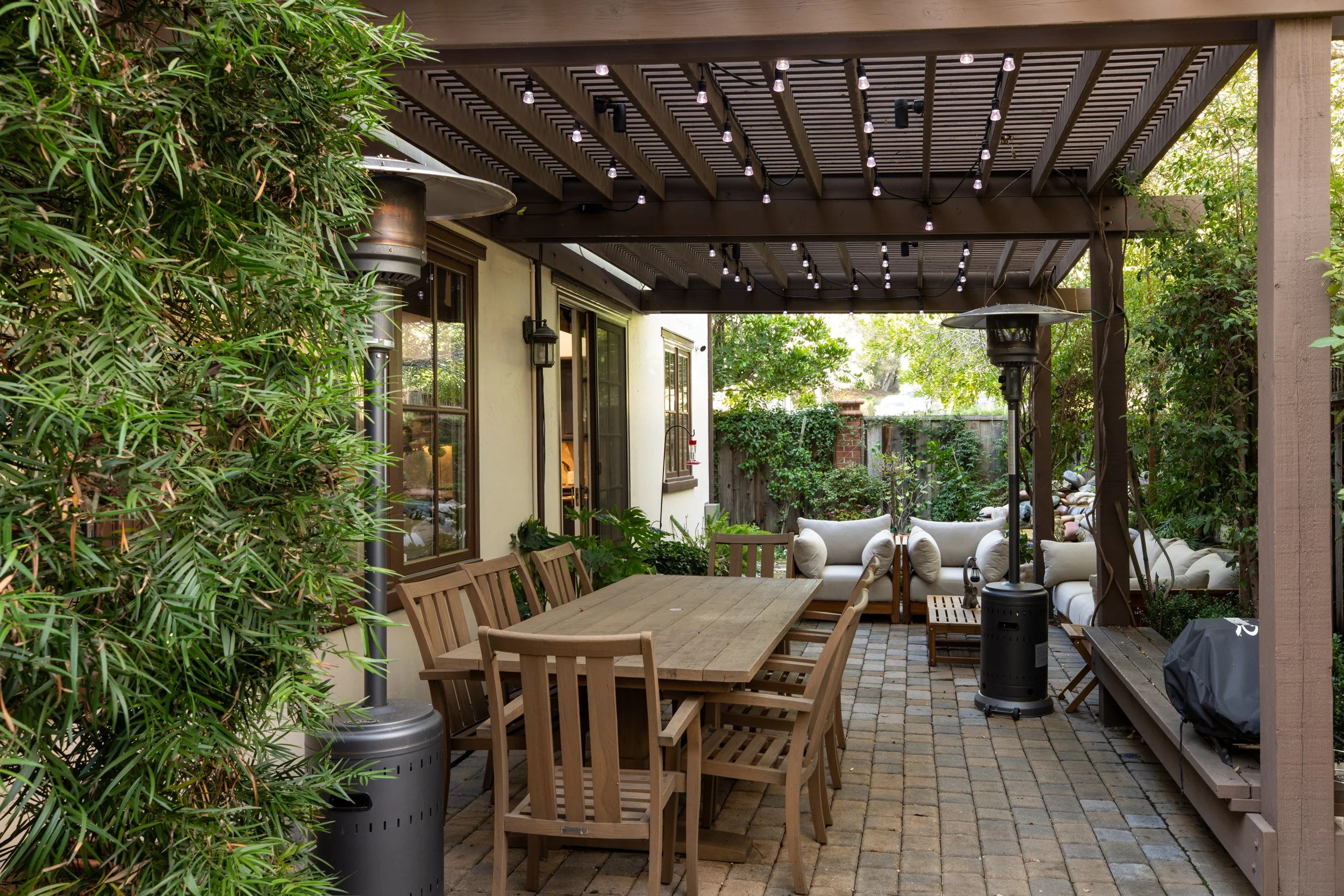 Outdoor patio with wooden dining table, chairs, and white cushioned sofas under a pergola with string lights, surrounded by greenery