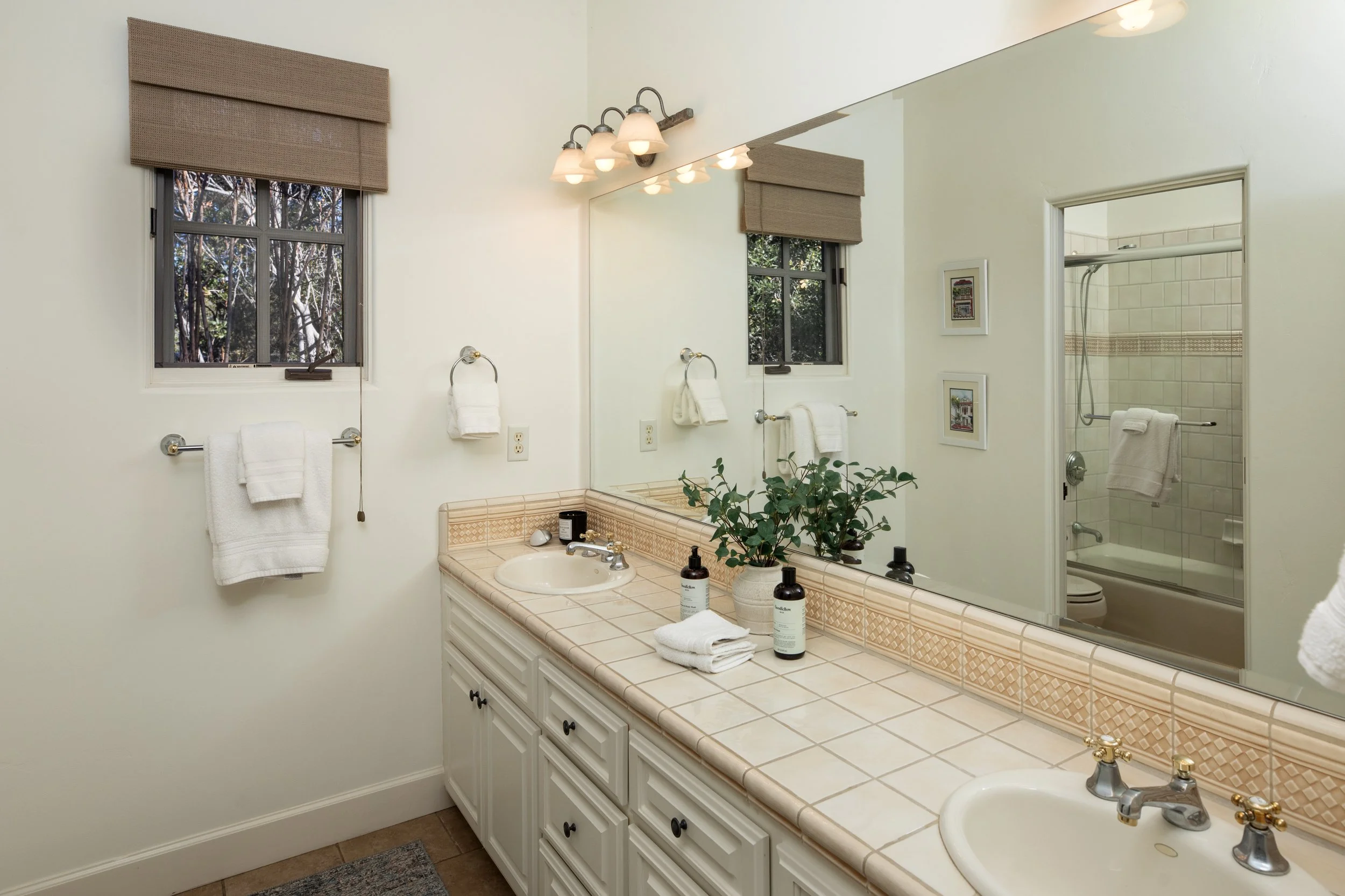 Bathroom with double sinks, large mirror, and window with brown window shades. There are towels on towel racks, plants on the counter, and a visible shower/bathtub in the background.