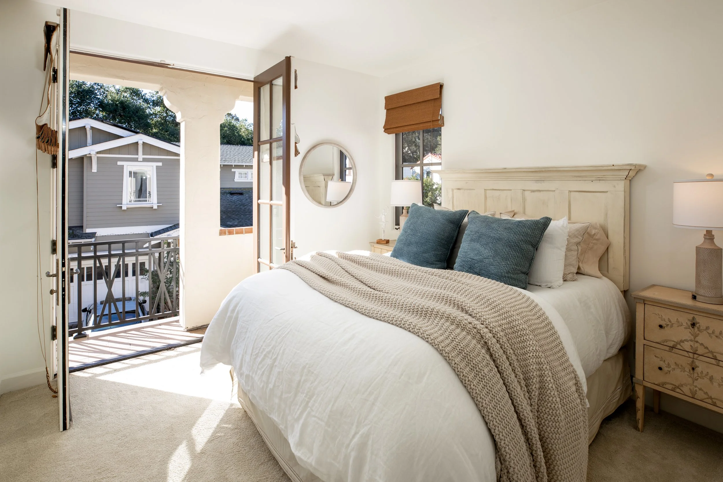 A bedroom with an open door leading to a small balcony. The room has a large bed with white bedding, two large blue pillows, a beige throw blanket, a wooden nightstand with a lamp, a round mirror on the wall, and a window with a brown shade. Sunlight