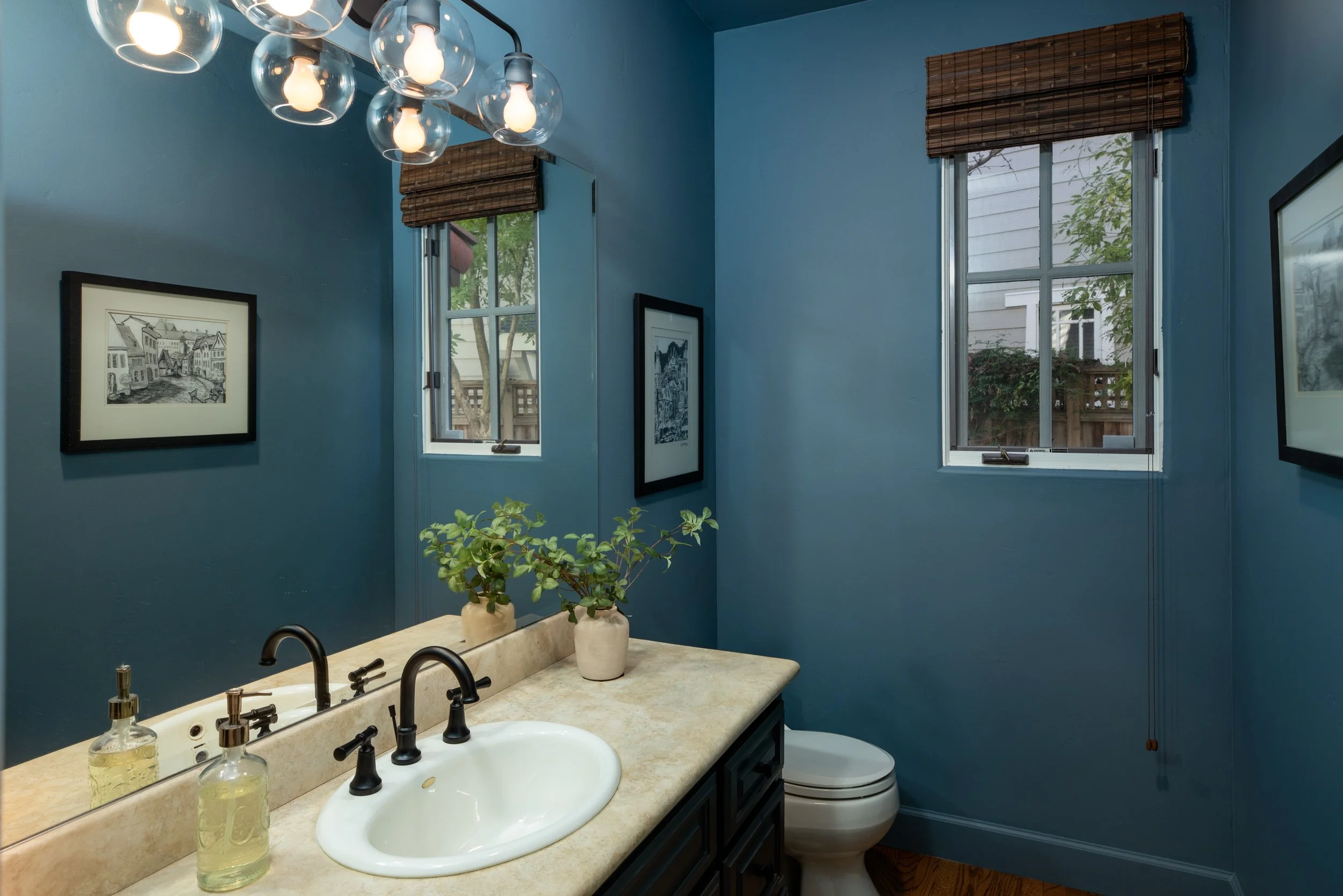 A bathroom with blue walls, a beige countertop with a white sink, two black faucets, and a mirror. On the counter, there are two soap dispensers and a small plant in a white pot. Two framed pictures hang on the walls, and there's a window with wooden