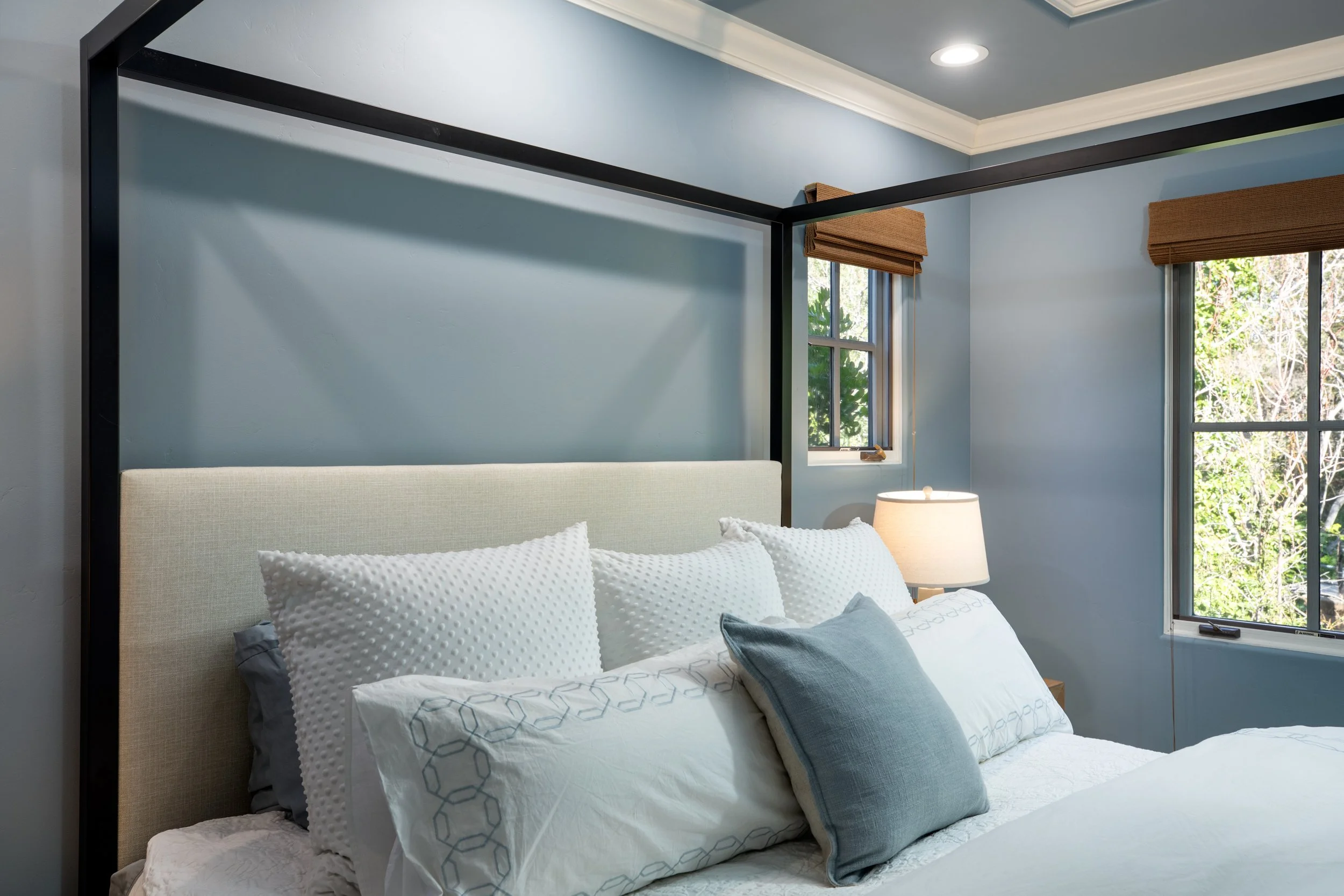 A bedroom with a light-colored bed, six pillows, a nightstand, a lamp, and windows showing greenery outside.