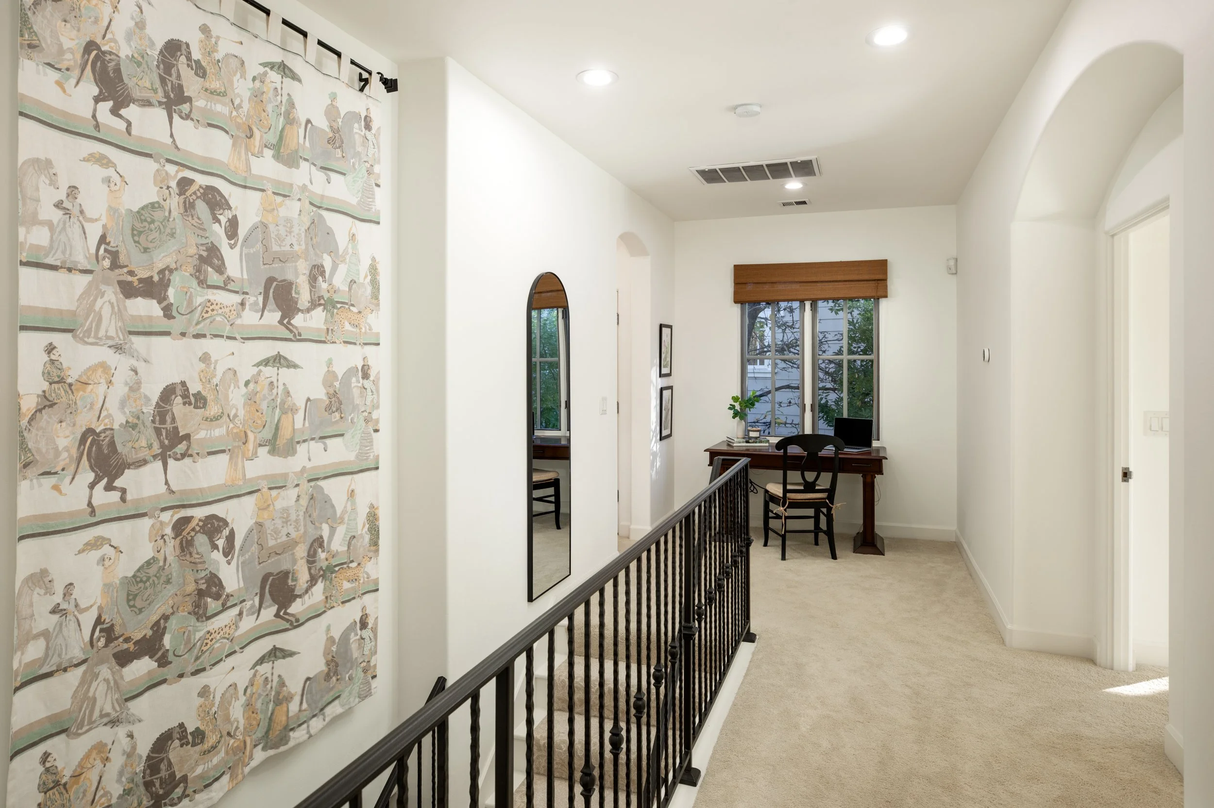 Interior view of a hallway with a black banister, a large tapestry on the wall depicting horses and people, a mirror, and a small desk with a chair near a window with wooden blinds.