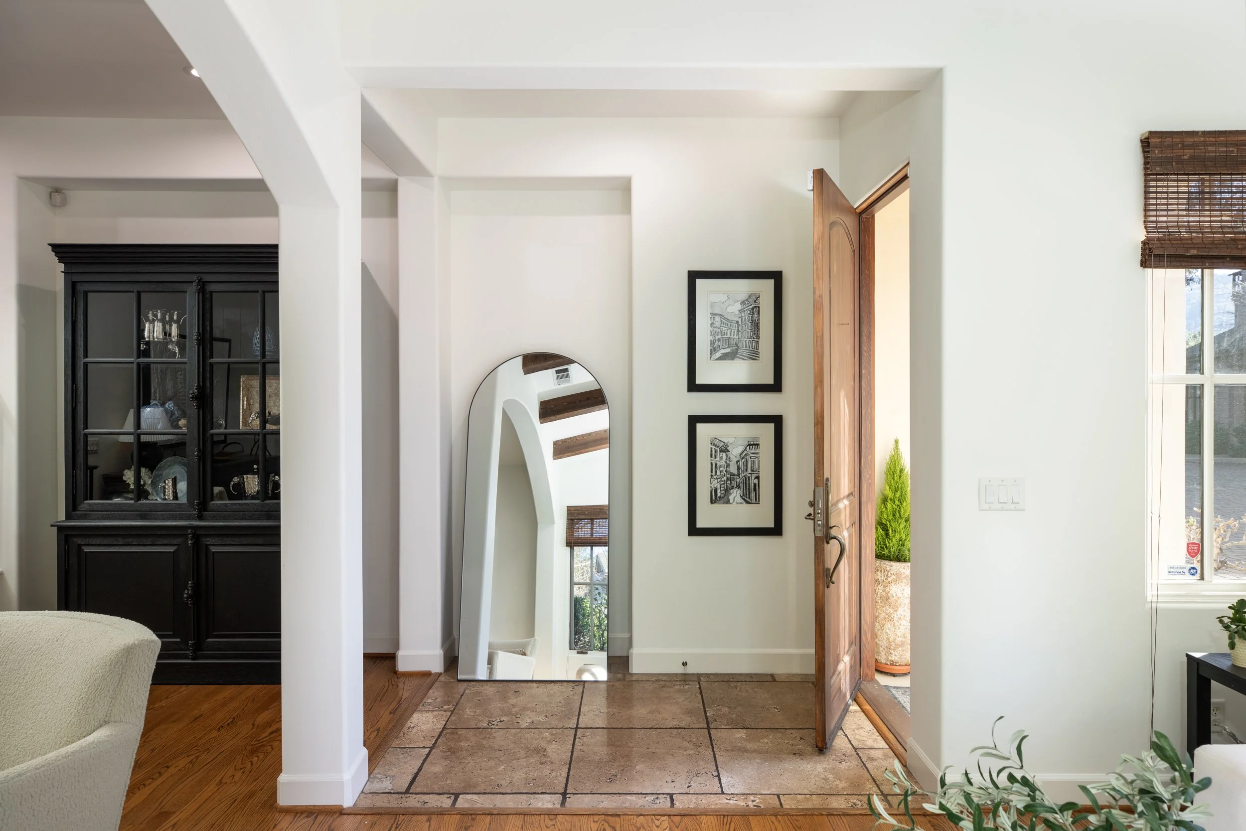 Entryway with open wooden door, a tall mirror leaning against the wall, two black-framed pictures, and a window with bamboo shade.