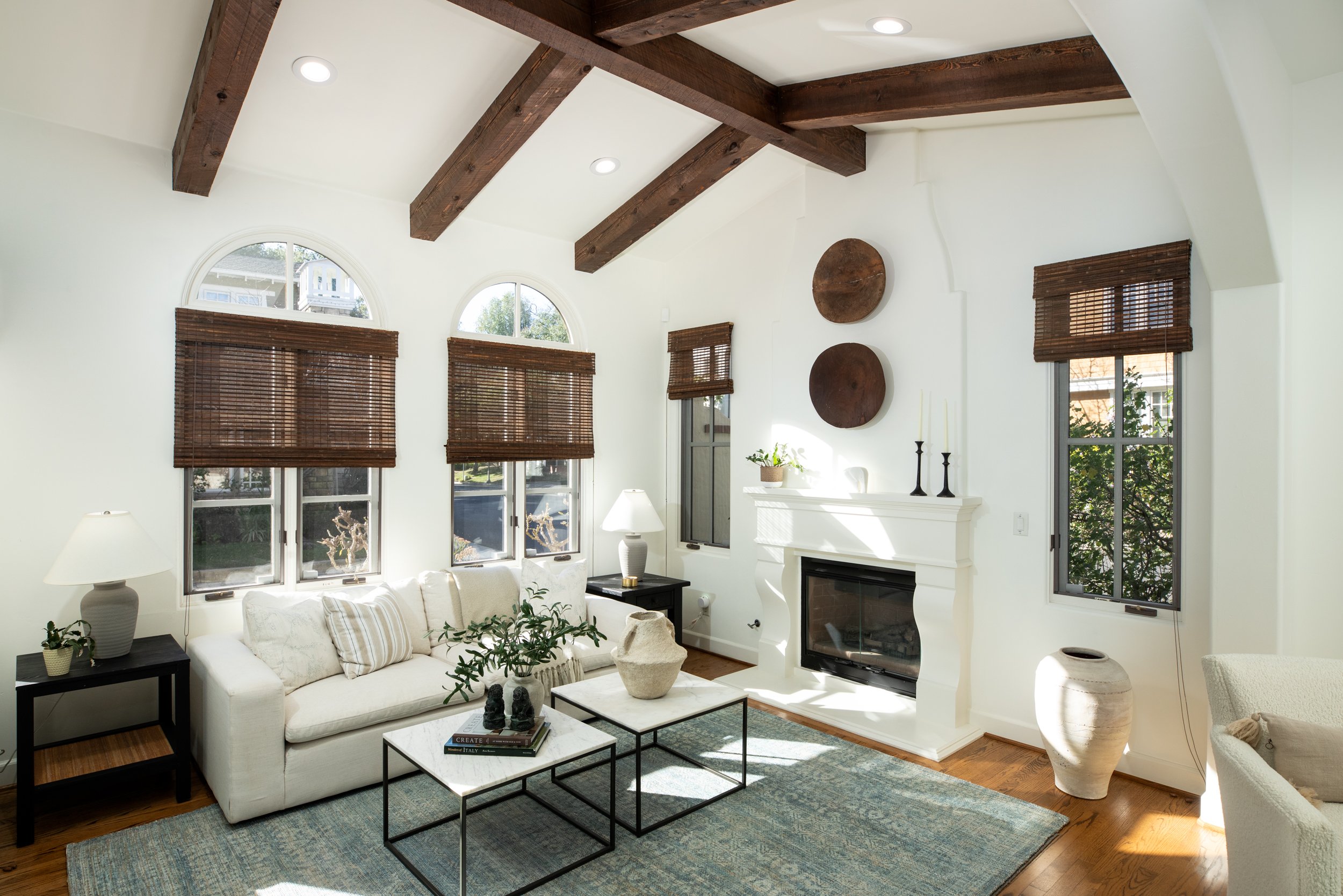 Bright living room with white walls, wooden ceiling beams, large arched windows with wooden blinds, white sofa with decorative pillows, black side tables with lamps, potted plants, woven basket, fireplace with white mantel, dark candle holders, abstr