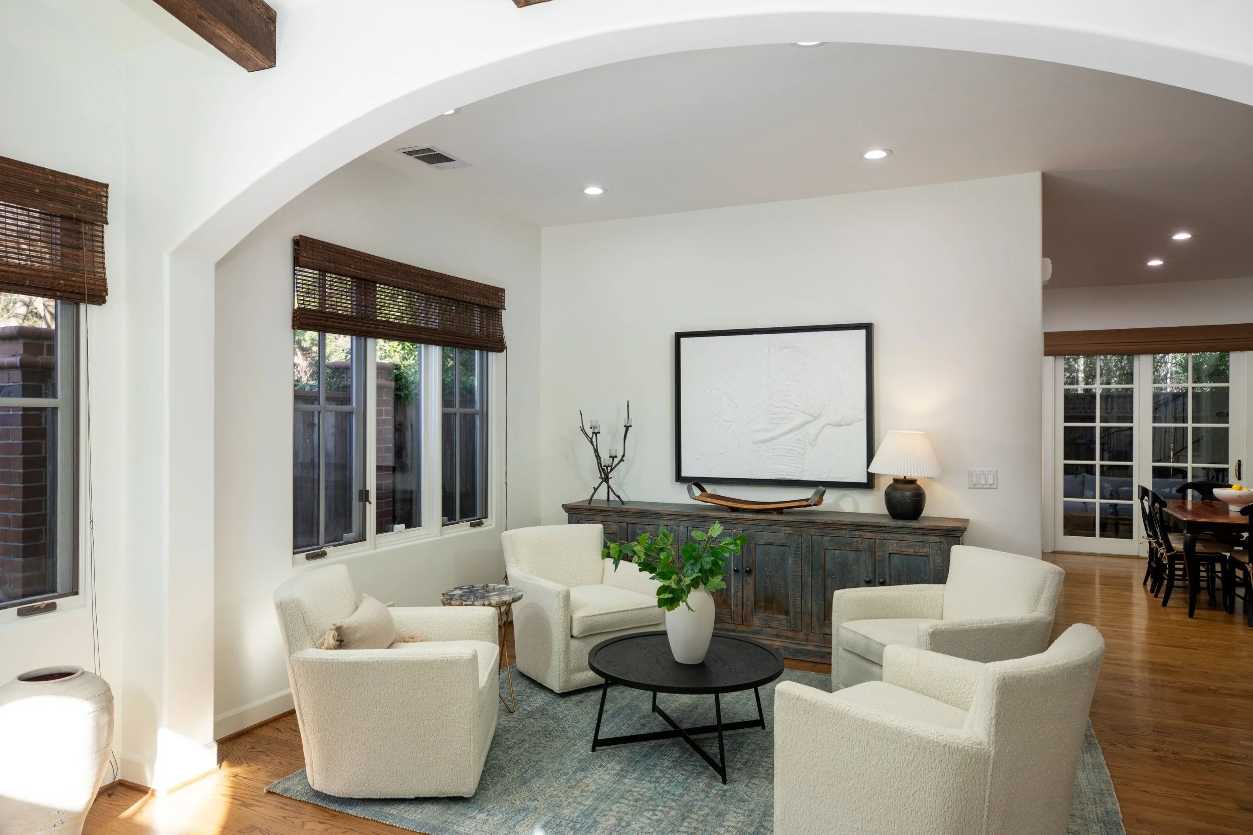 Living room with four white armchairs around a black coffee table with a vase of green leaves, a rustic cabinet with a lamp and art piece, and windows with brown blinds.
