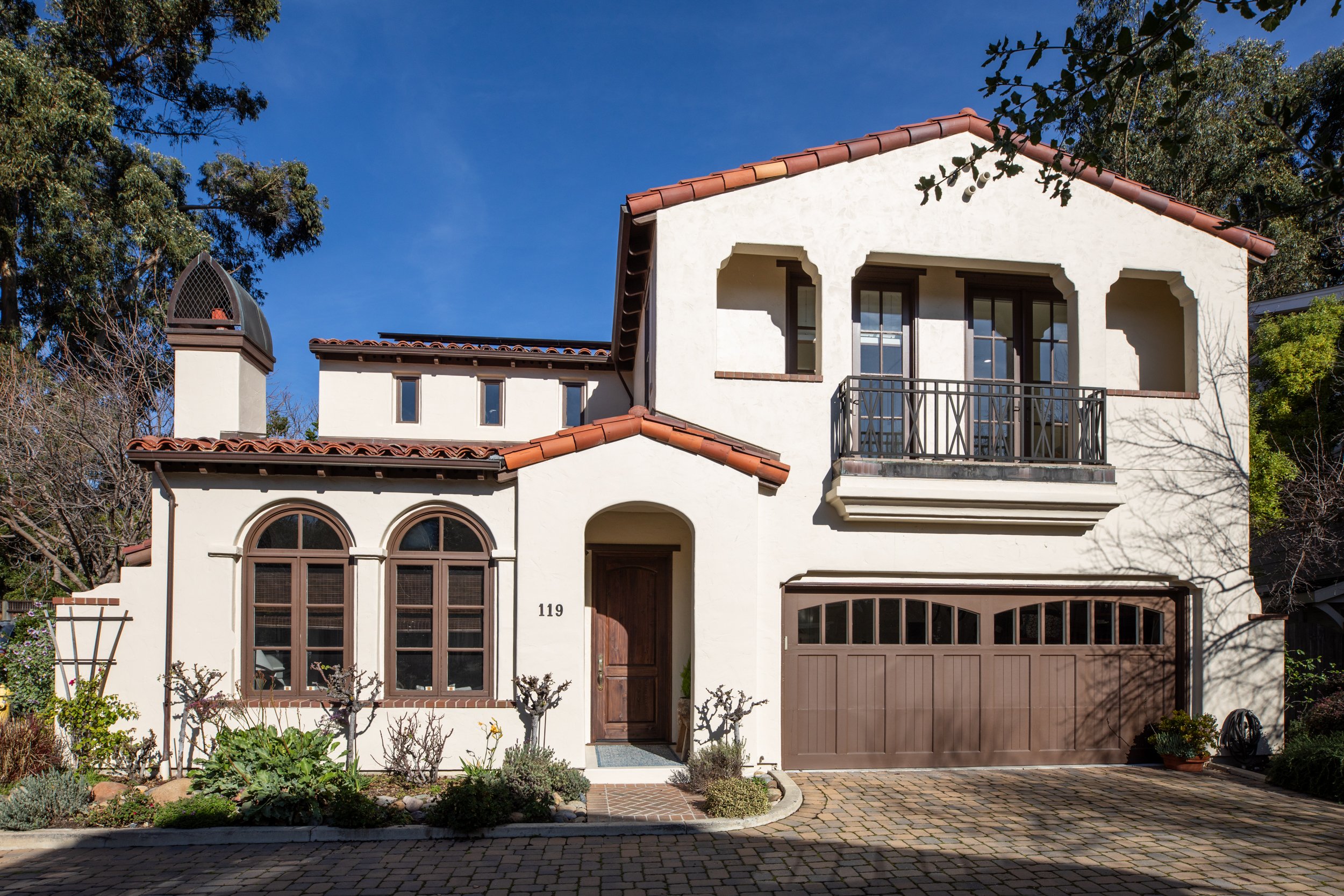 A two-story Mediterranean style house with white stucco walls, brown tiled roof, and arched windows. It has a front door, a two-car garage, and a small balcony above the garage, surrounded by trees and a small garden.