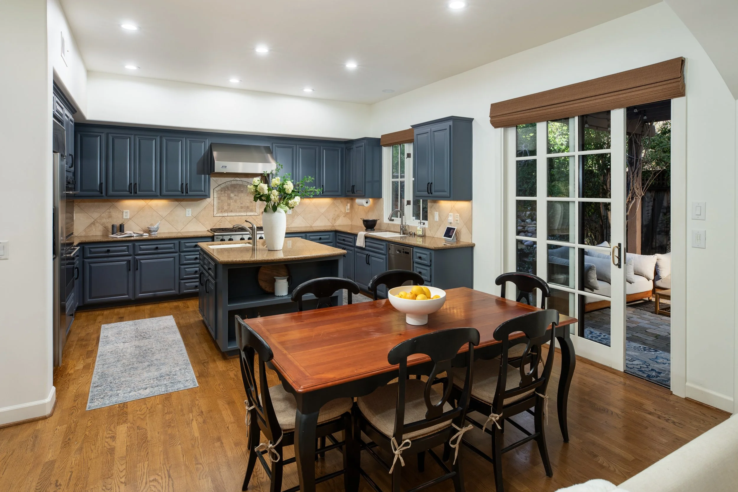 A kitchen with dark blue cabinets, beige tile backsplash, and a wooden dining table with six chairs. There are sliding glass doors leading to an outdoor patio with seating.